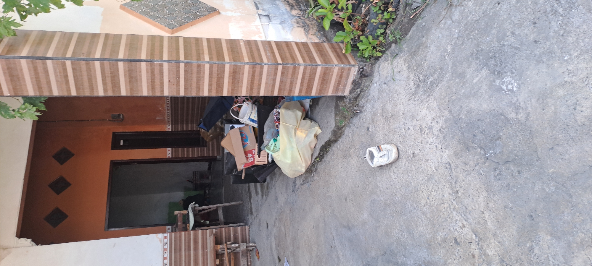 The image depicts an outdoor scene in front of a house, likely in Cemandi, Indonesia, characterized by a concrete ground and an entrance to a building. A single white sandal with orange and black stripes is placed on the concrete in the foreground. To the left of the sandal, a large, light-yellow plastic bag, possibly containing items, rests on the ground. Behind the plastic bag, a collection of cardboard boxes and a white bucket are visible, some stacked and others scattered. A dark blue bag and a black object are also present among the items. The entrance to the house features an orange-colored wall with decorative square indentations. A wooden stool and a table are positioned to the left of the entrance, near the house. A decorated pillar with horizontal stripes stands to the right of the entrance. In the background, part of the house's interior is visible, showing a dark doorway and a shadowy area. Green foliage can be seen to the upper right of the image, and a small cluster of green plants grows near the base of the pillar. The overall impression is one of an exterior space with discarded items and a humble dwelling.