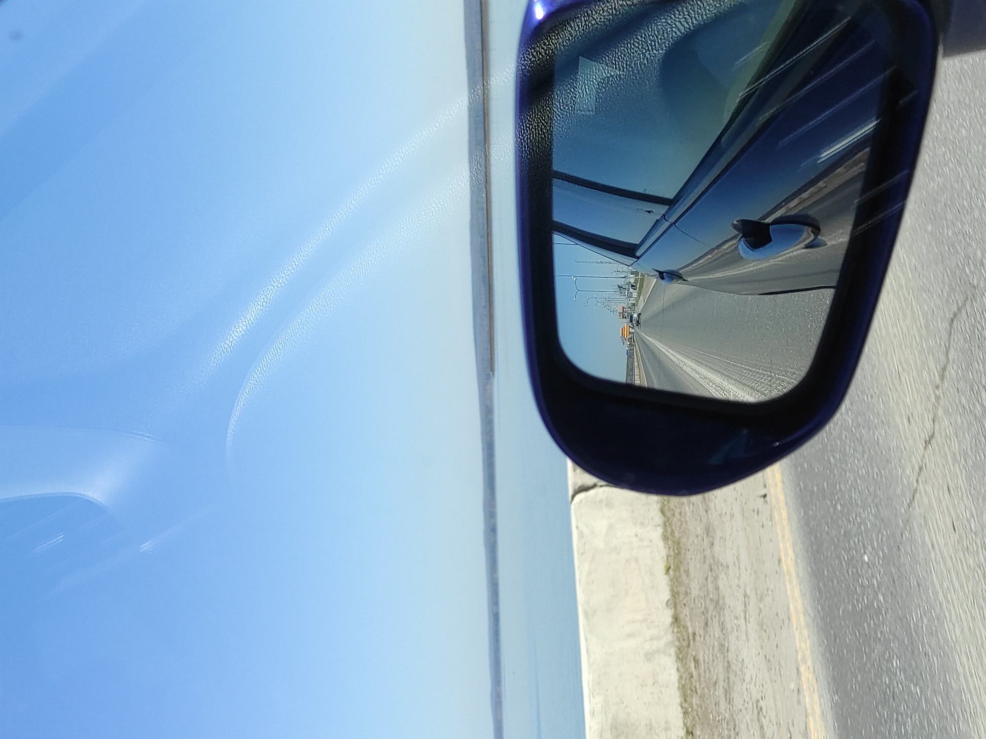 The image depicts a view from inside a moving vehicle, likely a car, showing the passenger side view and the side mirror. The mirror reflects the adjacent lane of the highway, displaying other vehicles, including cars and trucks, moving in the same direction. Streetlights are visible along the road. Outside the car window, the sky is blue and the water is still. The reflection on the window indicates that the vehicle is in motion. Based on the clues of the city name and water, this scene appears to be near the Kama River on the outskirts of the city of Chaikovsky, Russia.