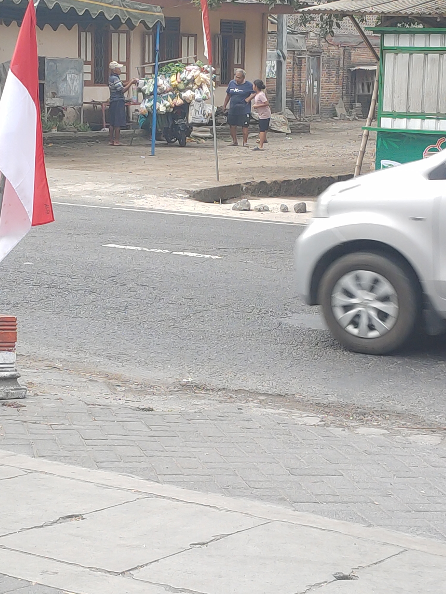 This is a full-frame, eye-level shot of a street scene in Kanigoro, Indonesia. In the foreground, a silver car is partially visible, with its front wheel and fender in motion. To the left, a red and white Indonesian flag on a pole stands prominently. Behind the flag, a man in a light-colored hat and dark shorts is standing next to a motorcycle laden with goods in plastic bags. He appears to be attending to something on the motorcycle. Further back, a man in a dark blue t-shirt and shorts and a younger person in a light pink shirt and dark shorts are standing near a brick building. In the background, there is a simple green and white kiosk structure. The ground is a mix of asphalt, dirt, and paved brick. The overall environment suggests a commercial or market area.