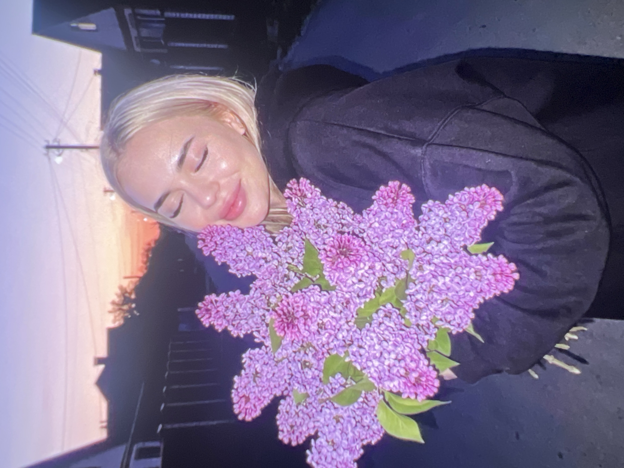 A person is captured in a close-up, rotated image, embracing a large bouquet of purple and pink lilac flowers. The subject, a woman with blonde hair, has her eyes gently closed and a soft, contented smile on her face, resting her cheek against the blossoms. Her skin appears luminous, reflecting the warm light. She is wearing a dark, possibly black, hooded sweatshirt.
The setting appears to be outdoors during either sunrise or sunset, indicated by the gradient sky in the background transitioning from a soft light purple to a warm orange-pink hue. Silhouetted structures, resembling houses or buildings, and utility poles with power lines are visible against the colorful sky, suggesting a residential or urban environment. The overall ambiance is peaceful and joyful, conveying a moment of appreciation for nature. No text is visible in the image.