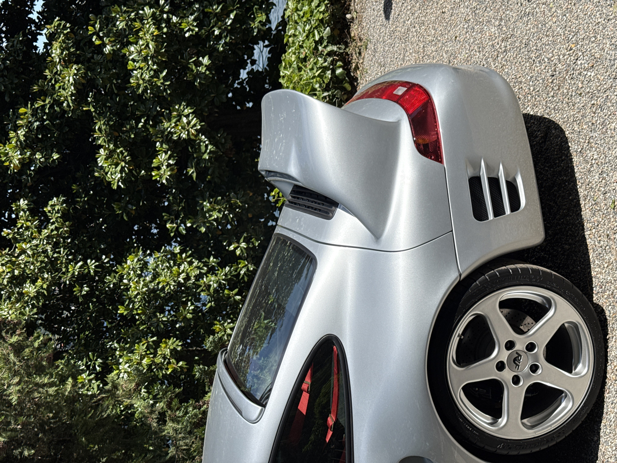 The image shows the rear of a silver Porsche, possibly a 911 or similar model, with a large rear wing. The car is parked on a gravel surface. The background includes lush green foliage, suggesting a location near trees or a park. The location is Como, Italy. The car appears to be well-maintained and possibly modified, judging by the aftermarket wheel and rear wing. The taillights are red, and the rear vents are visible. Inside the car, red seat belts are visible. The scene is outdoors in daylight.