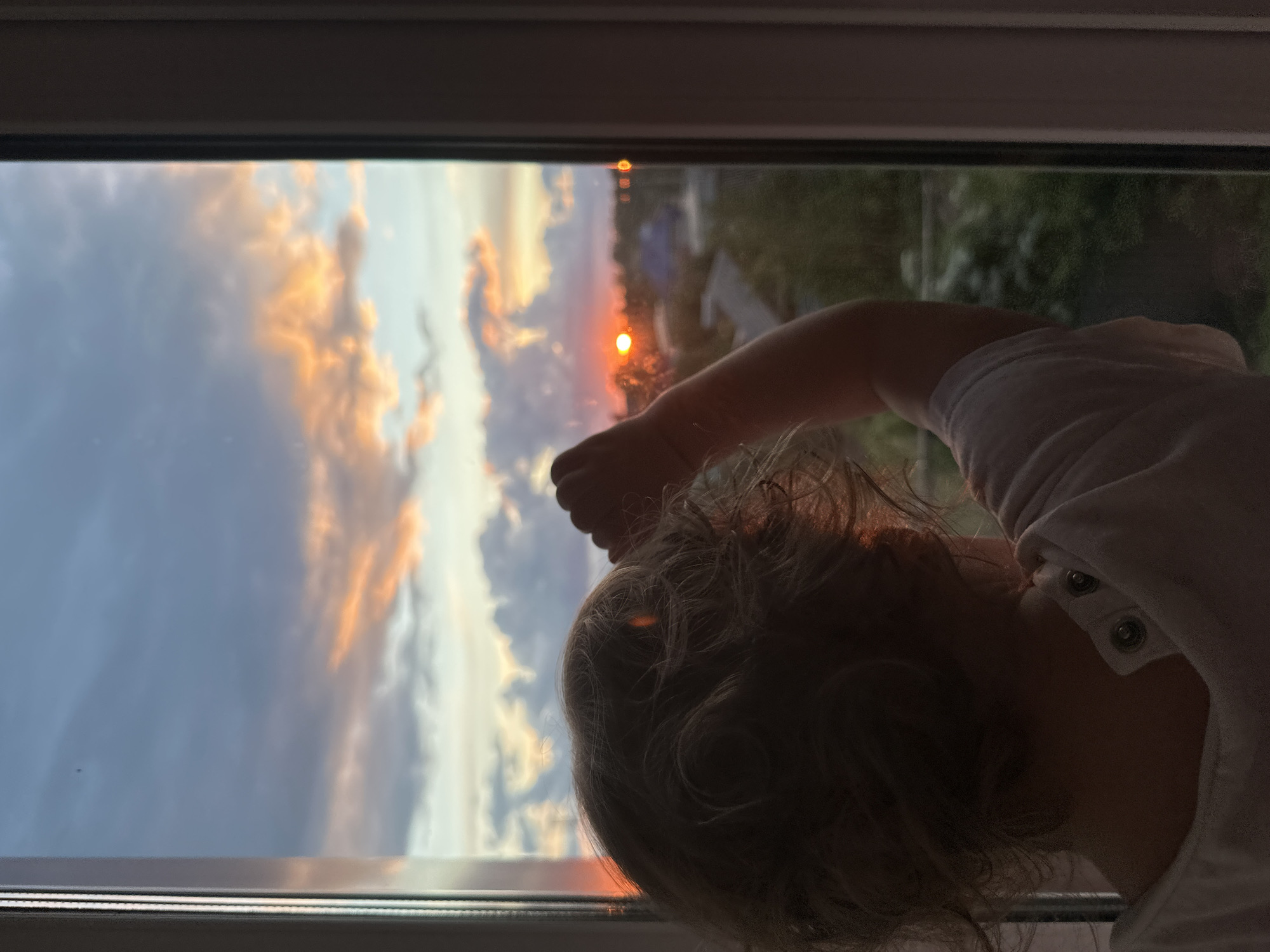 A child is gazing out of a window at a sunset, in Unknown, Unknown. The child, wearing a white top, has their hand near the window. Outside, the sky is a blend of blue, orange, and gray clouds.