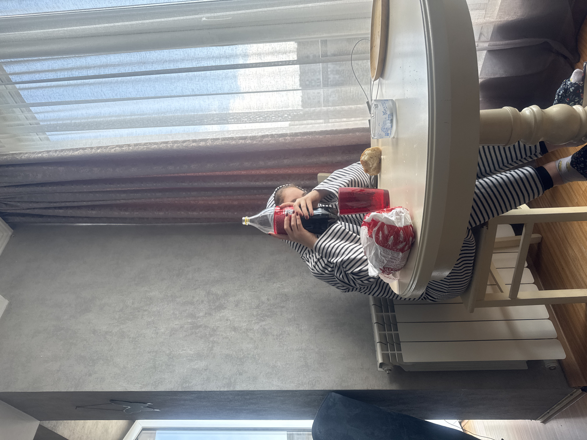 A person is seated at a white round table near a window in a room. The person is wearing a striped shirt and pants. They are holding a large Coca-Cola bottle with both hands. A red box, white bag, a piece of bread, and a clear glass are on the table. The room has a gray wall, a radiator, and curtains covering the window. Outside the window, there is a view of the city, likely Tbilisi, Georgia.