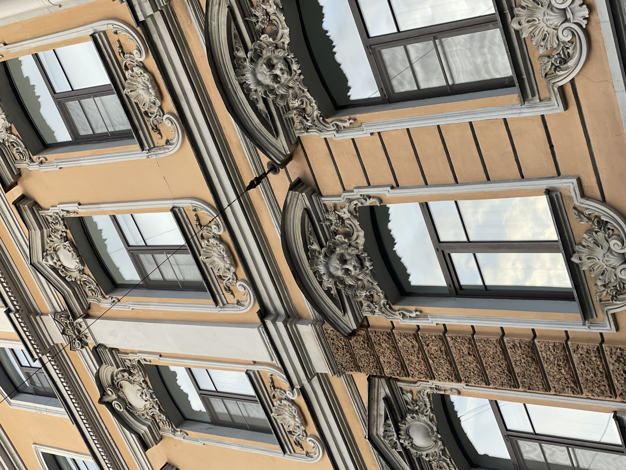 The image shows a close-up, low-angle view of a building facade in Saint Petersburg, Russia. The facade is characterized by ornate Art Nouveau detailing, including decorative window frames with floral and grotesque face motifs. The building's exterior is painted in light yellow and beige tones, with contrasting white and grey trim highlighting the architectural details. Rectangular windows with dark frames reflect the sky. Electrical wires are visible against the facade. The overall impression is of historical architecture with rich ornamentation.
