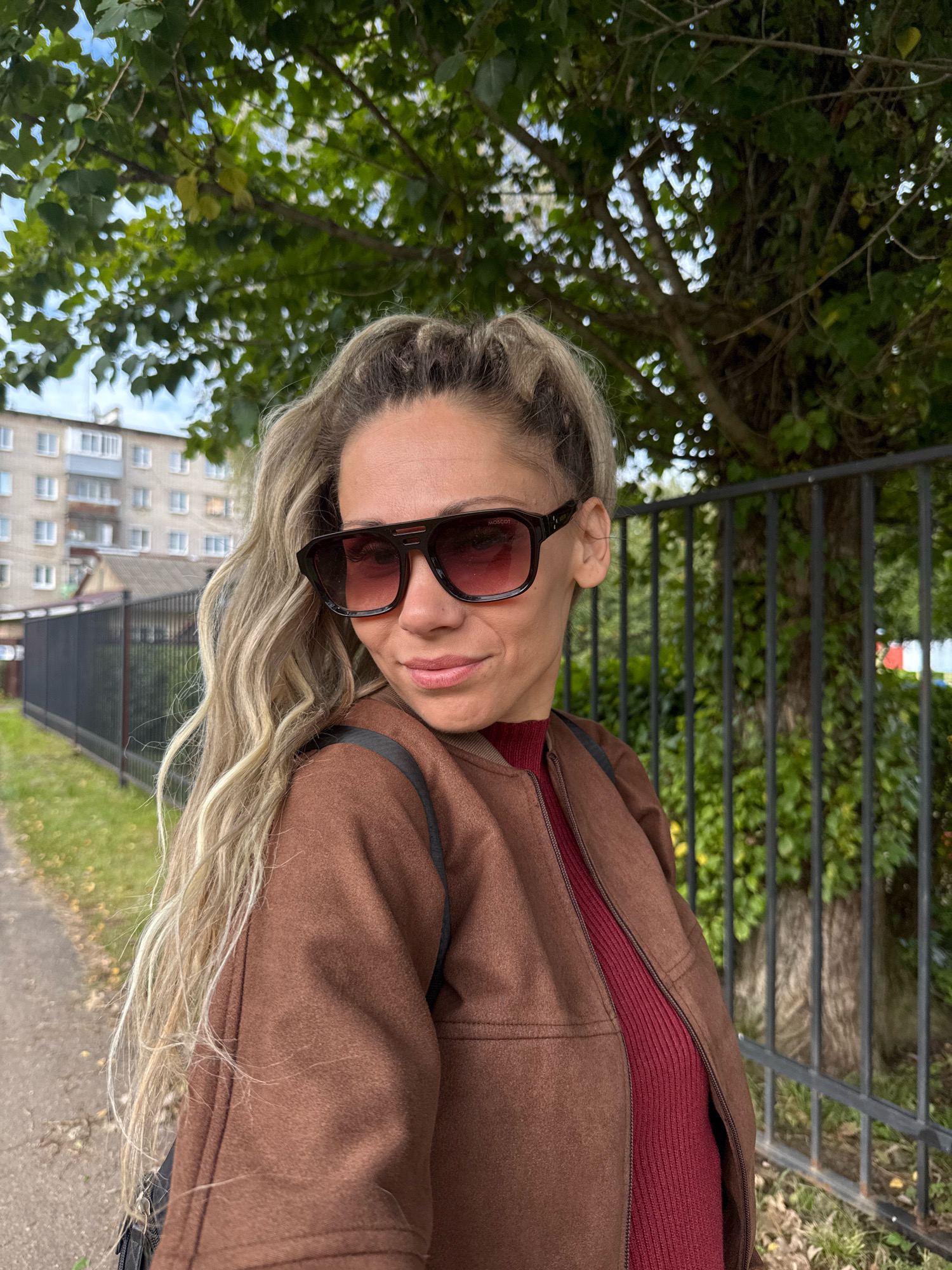 A medium shot captures a woman with blonde, braided hair, wearing large, dark sunglasses and a brown jacket over a maroon top. She is positioned outdoors in front of a black metal fence with a tree and foliage visible behind her. To the left, a sidewalk, grass, and a multi-story building are present. The scene appears to be a residential street in the city of Zhukovo, Russia, under an overcast sky.