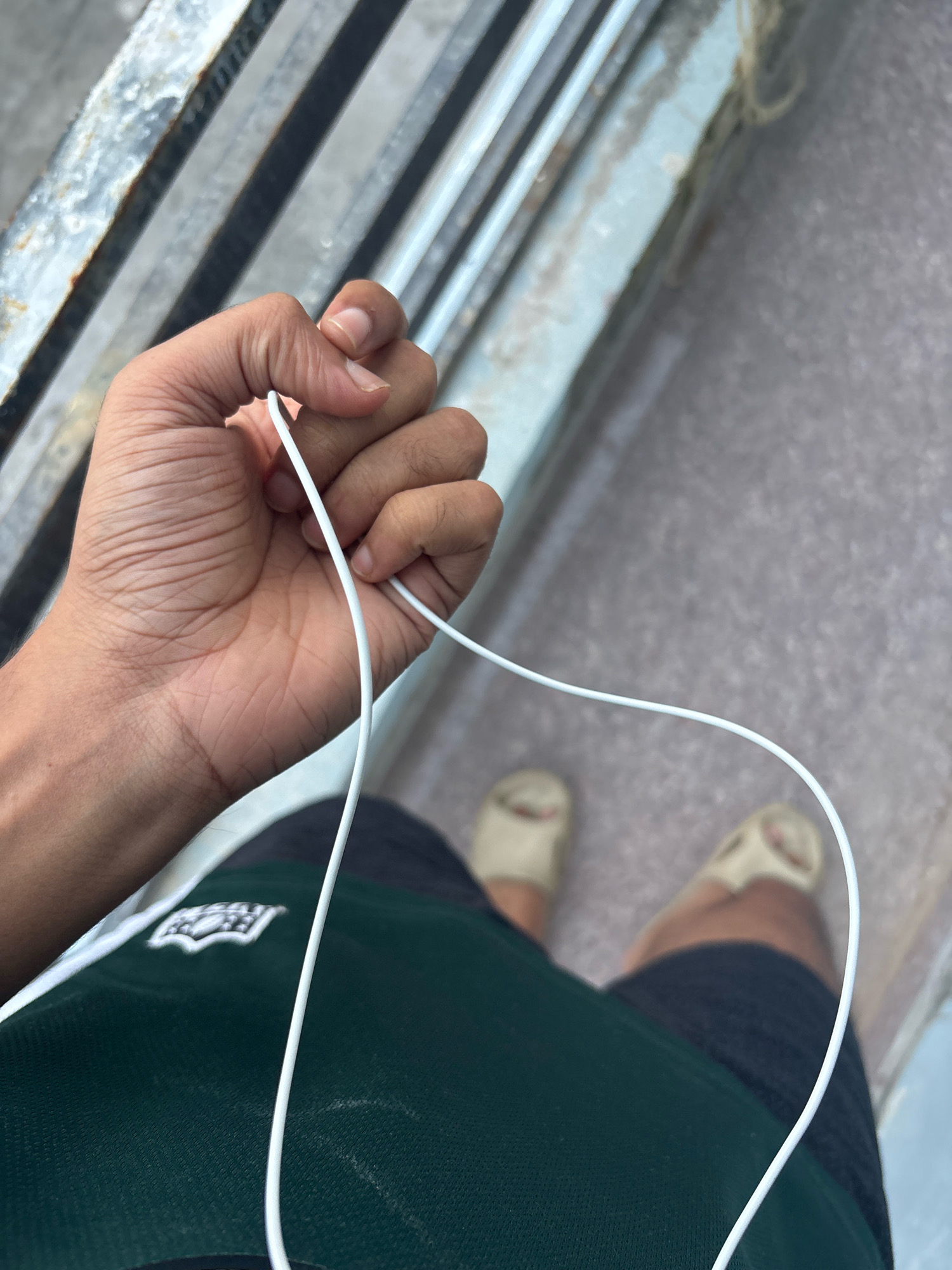 A close-up shot from a first-person perspective shows a hand holding a white cable, likely from earbuds. The hand is clenched around the cable, with fingers bent and knuckles visible. The skin of the hand shows creases and some hair. The cable forms a loop in front of the hand. In the background, out of focus, are a person's legs wearing dark shorts and light-colored slip-on shoes. The ground appears to be paved. To the left, there is a metal railing with a weathered, possibly rusty, texture, suggesting an outdoor environment, possibly near a street or elevated walkway. The overall lighting suggests it is daytime. This scene could be in the city of Bhatta, India, given the mention, though no specific landmarks confirm this location.