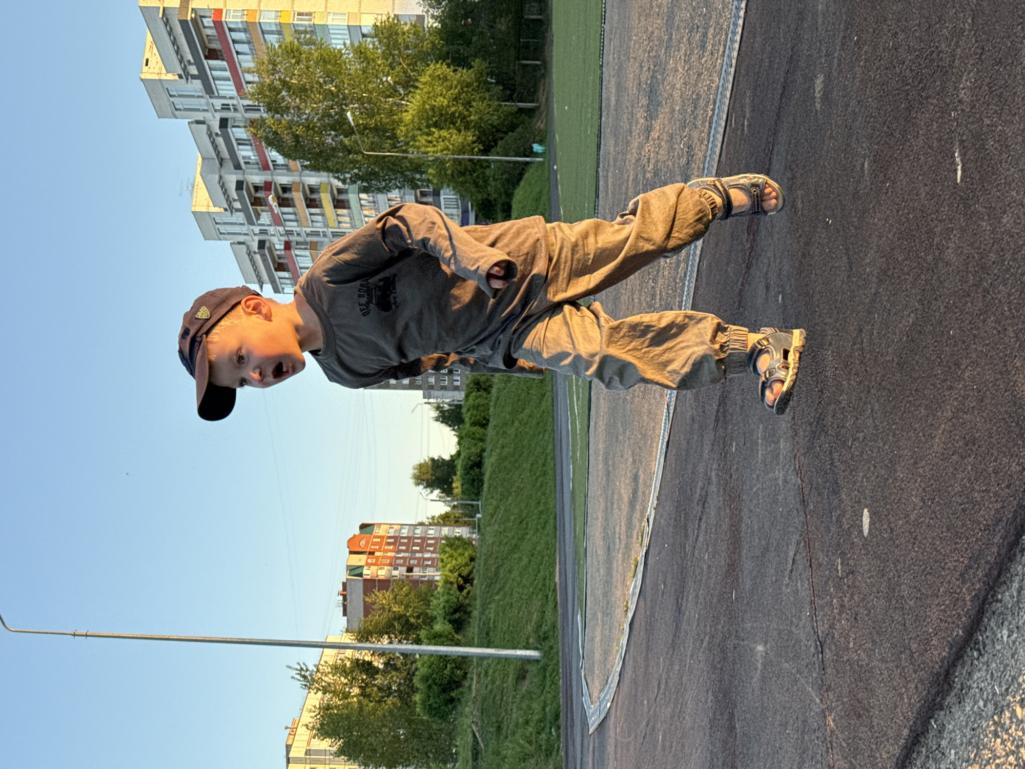 A boy is standing on a dark paved surface in the city of Chaikovsky, Russia. He is wearing a dark baseball cap, a long-sleeved gray shirt, gray pants, and sandals. The background includes apartment buildings, trees, grass, and a blue sky. A metal light pole is visible on the left. The pavement appears to be part of a playground or recreational area.