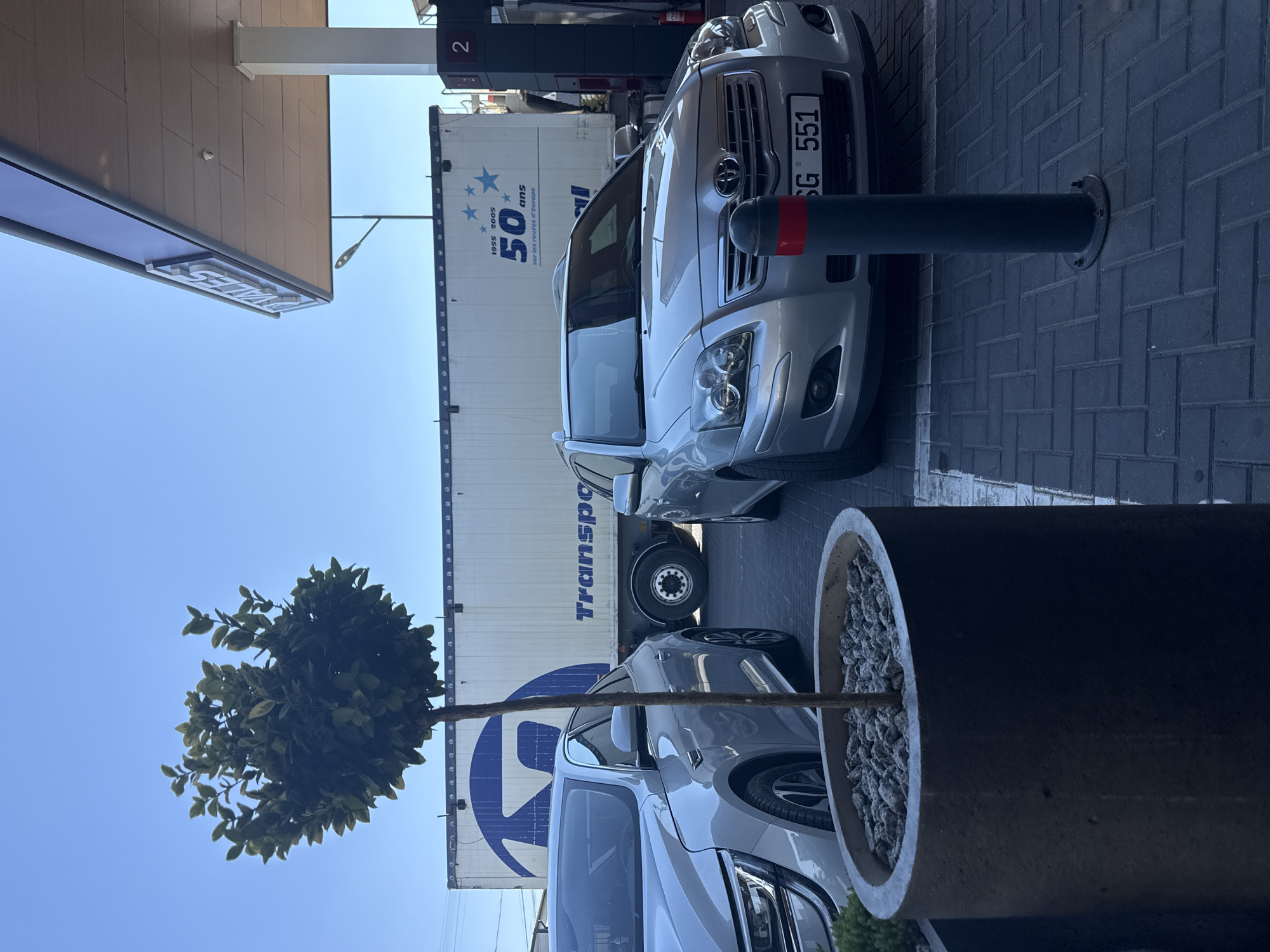 The image depicts a street scene in Comrat, Moldova, under a clear blue sky. In the foreground, a silver sedan is parked on a brick-patterned surface. Behind it, a truck with a white trailer is visible, featuring blue lettering that says "Transport" and "50 ans sur les routes d'Europe" along with blue stars. To the right, a gas station canopy with white signage and a white pillar extends over the parking area. Another silver car is partially visible on the far right. A tall, thin tree in a circular concrete planter stands in the left foreground. A dark bollard with a red stripe is positioned in front of the silver sedan.