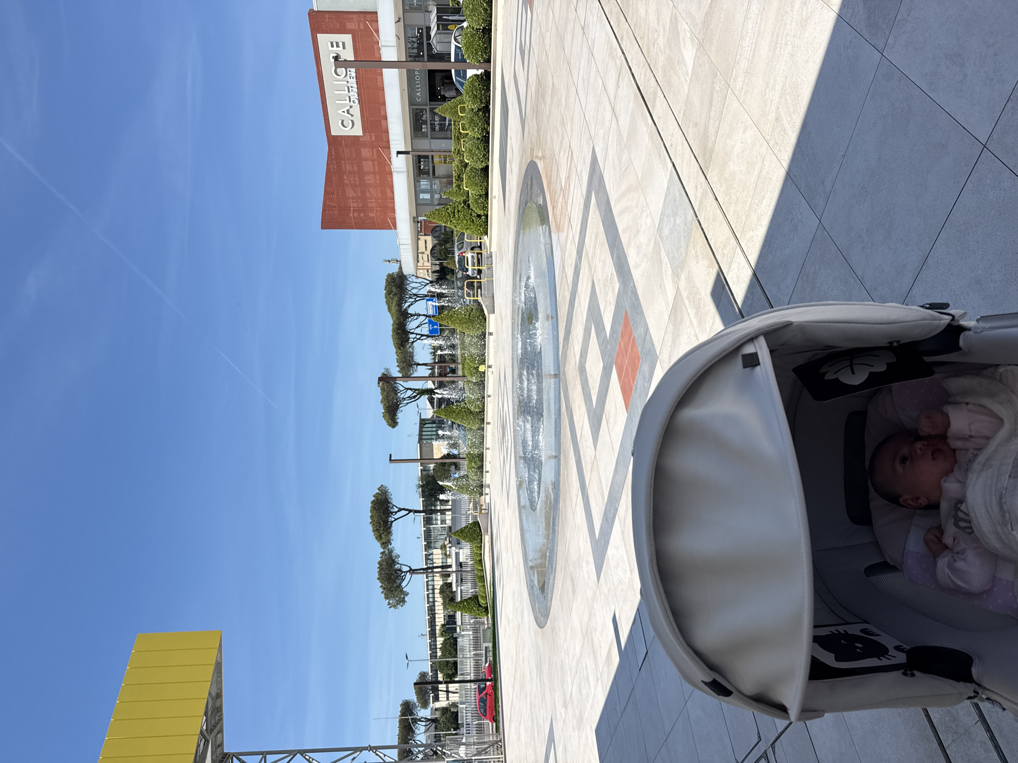 The image depicts an outdoor scene in Pomezia, Italy. A baby is lying in a stroller, positioned on a paved walkway. Adjacent to the walkway is a decorative fountain and a row of trees. Buildings are visible in the background, including a structure with the word "CALLIOPE" displayed. The sky is clear and blue. Construction scaffolding is visible on the left side of the frame.