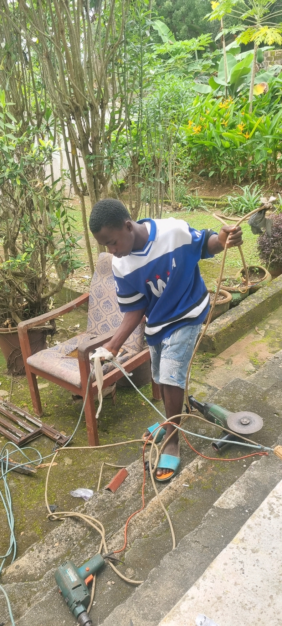 In Iju, Nigeria, a young Black male is pictured outdoors, appearing to be engaged in some form of repair or construction work. He is wearing a blue and white t-shirt with a V-neck and a logo on the left side of his chest, along with light-colored denim shorts and blue flip-flops. The young man is holding a coiled rope or cable in his right hand and a piece of white material in his left hand, which he is interacting with, possibly preparing it for use.
He is standing on a set of concrete steps, partially surrounded by overgrown vegetation and trees. To his left, an old, worn armchair with a patterned fabric is positioned. Various tools and electrical cords are scattered around the steps and the ground. Visible items include a green angle grinder to his right, and a green electric drill with a chuck on the steps below him. Some metal framing is also present on the ground to the left of the armchair. The environment suggests a somewhat unkempt outdoor area, possibly a backyard or yard space, with elements of both nature and discarded materials. The lighting indicates it is daytime.