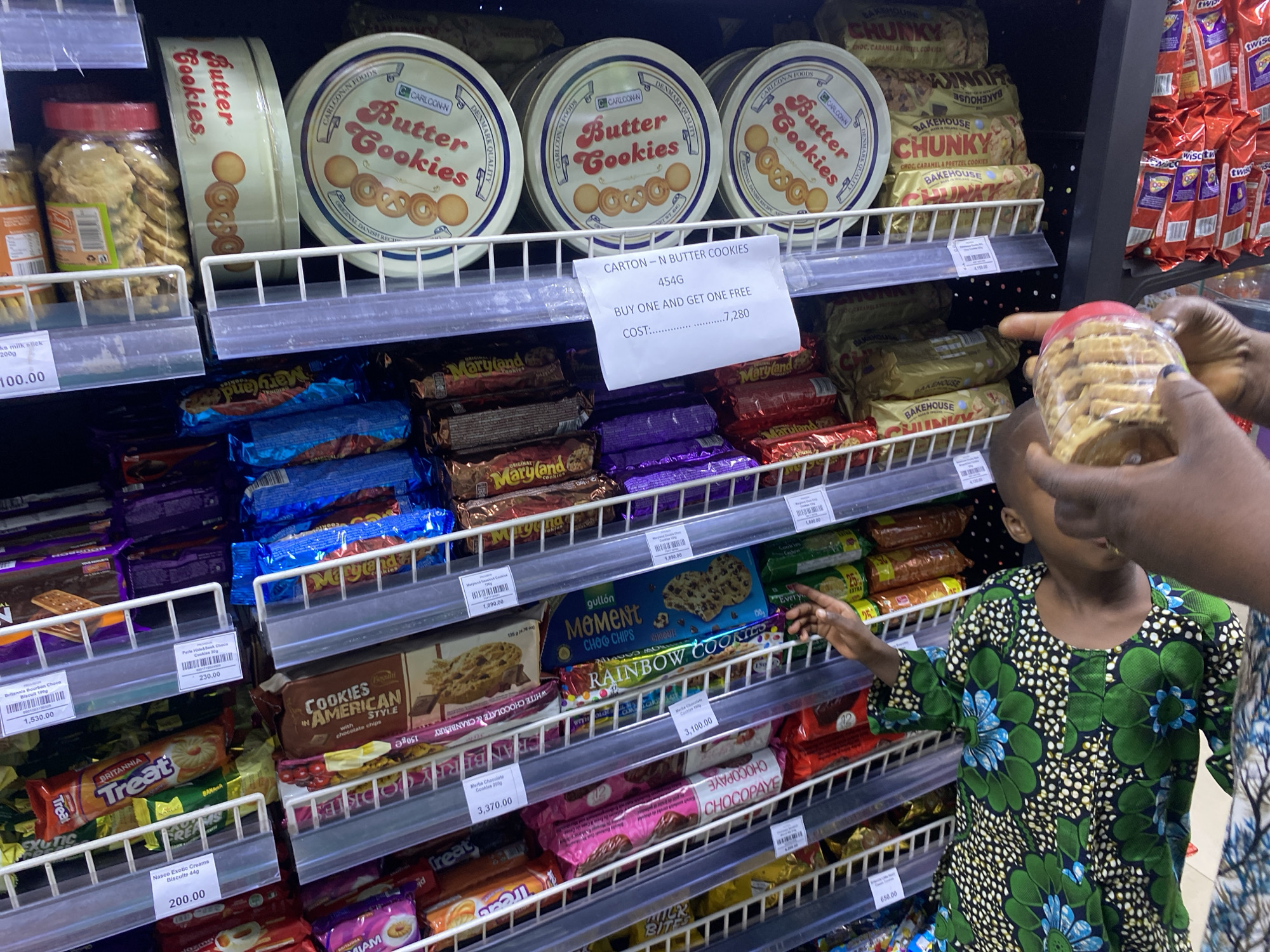 The image shows an interior shot of a shop or supermarket in Lagos, Nigeria. The focus is on a section of shelving stocked with various brands of cookies and biscuits. A young person in a green and black floral patterned shirt is visible, seemingly reaching for a pack of "Rainbow Cookies" from the "gullón Moment" brand. An adult's hand is visible near the top right, holding a jar of cookies. The shelves are lined with products such as "Carlson-n Butter Cookies" in tins, "Maryland" cookies, "Britannia Treat" biscuits, "Cookies in American style", and "Merba Chocolate Cookies". Prices are displayed for many of the products. A sign above the "Carlson-n Butter Cookies" states "Buy one and get one free" and lists the cost as 7,280. The setting appears to be a well-lit retail environment.