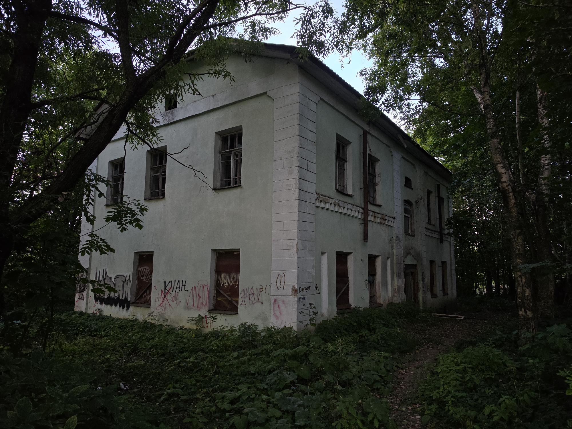 The image shows an abandoned two-story building in Strelna, Russia, overgrown with vegetation. The building's pale green facade is covered in graffiti. Most windows are boarded up with wooden planks. The structure appears to be in disrepair, with crumbling plaster and missing architectural details. Tall trees with dense foliage surround the building, casting deep shadows and obscuring parts of the structure and the surrounding area. A narrow, unpaved path leads away from the building into the woods. The overall atmosphere is one of decay and neglect.