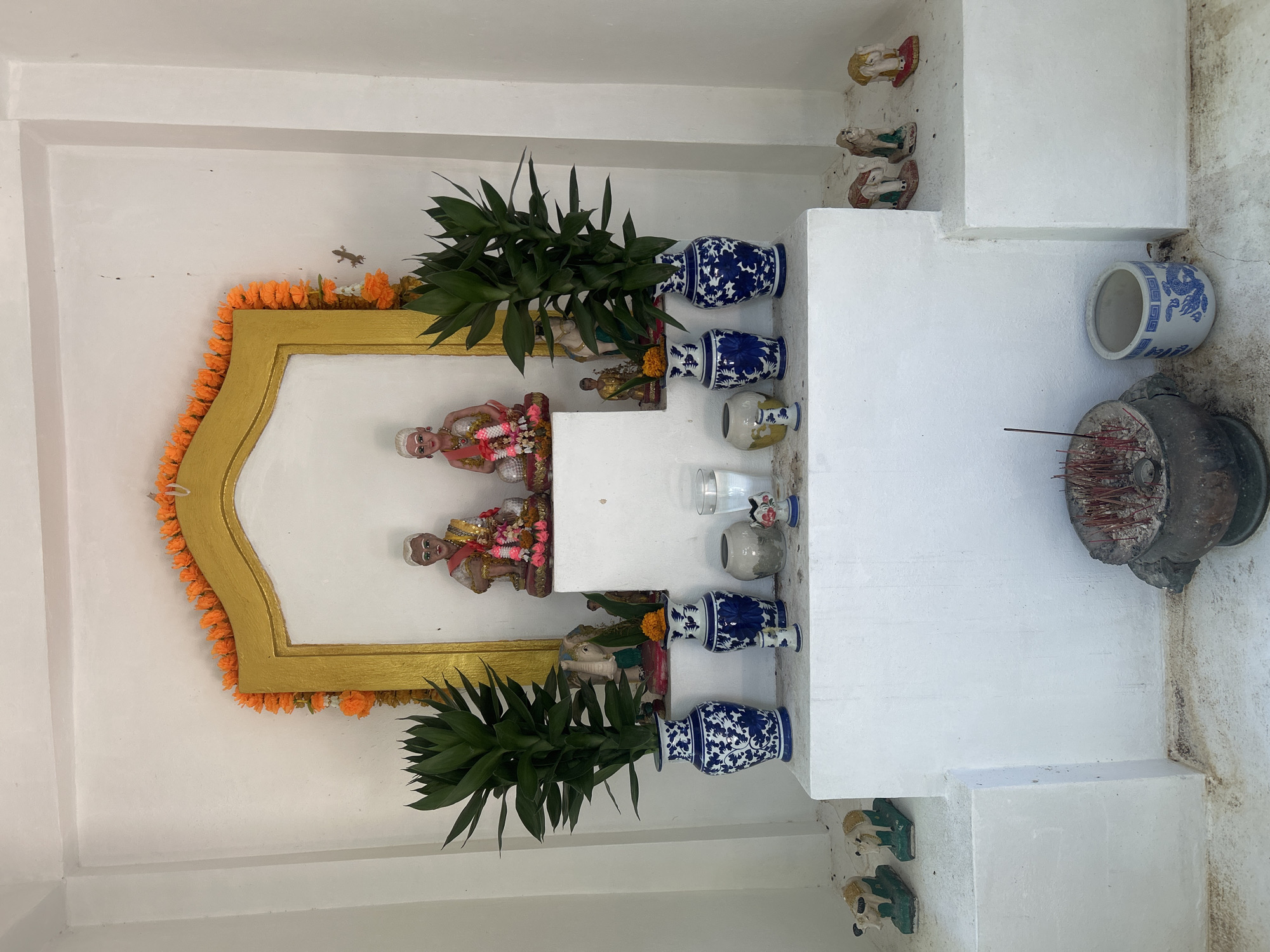 This is a head-on, medium shot of a small, white, shrine-like structure set against a white wall, likely in ตำบลเกาะพะงัน, Thailand. The shrine is decorated with a gold, arched frame adorned with a garland of small orange flowers. At the top of the shrine, two small, seated figures, likely religious icons, are positioned centrally. They are depicted with pale skin, light-colored hair, and wear colorful robes, adorned with floral garlands. Flanking these figures are tall, leafy green plants in blue and white patterned ceramic vases.
Below the main figures, on the first tiered level of the shrine, are various decorative objects. These include a small, white ceramic pot, a glass of water, and a few other smaller decorative items. To the left of the central figures, on a slightly lower ledge, is a ceramic elephant figurine. To the right, another similar elephant figurine is visible.
The second tiered level of the shrine, lower and extending forward, features more decorative elements. On the left, two small animal figurines, possibly cows or oxen, are placed on green bases. On the right, three more small animal figurines, also resembling cows or oxen with different colored bases, are visible.
At the very front of the shrine, on the ground, is a large, dark, weathered metal urn filled with incense sticks, some of which are lit and smoking. Next to the urn is a small, white and blue ceramic pot with a dragon motif. The ground surface is a light-colored, somewhat textured material, with some visible dirt and debris. The lighting appears to be natural daylight, casting soft shadows.