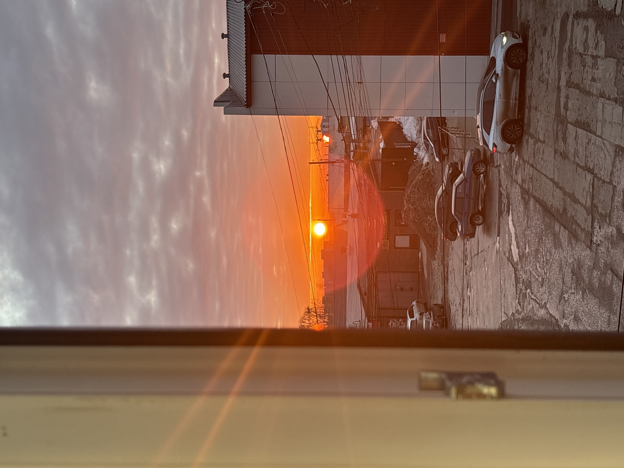 The image depicts an urban-industrial landscape in Chaikovsky, Russia, viewed from a window at either sunrise or sunset, characterized by a vibrant orange sky. A light-colored window frame, exhibiting distinct sun flares, occupies the left portion of the scene. The sky features a low-lying, intense orange sun, transitioning to hues of red, grey, and pink in the scattered clouds above.
Below the horizon, a commercial or industrial area is visible. On the right, a multi-story building with a facade split between white and dark brown sections, topped by a ridged roof, is prominent. Numerous overhead power lines are strung in front of this structure. Further into the mid-ground and to the left, several single-story industrial buildings, constructed of brick with large metal roll-up doors, are present. One building features a sign with Cyrillic script, including the term 'АВТО' (auto).
The ground consists of a paved lot, which appears wet and possibly icy, displaying visible puddles and irregular patterns. Patches of dirty snow are scattered across the lot, and a substantial pile of dark material, potentially earth or debris, is visible in the mid-left. Several vehicles are parked on the lot: a silver or white SUV (resembling a Volkswagen ID.4) is positioned in the lower right foreground. Other parked vehicles include a dark sedan, a blue or grey older model sedan, and a white SUV in various locations within the scene. Bare trees are discernible on the distant horizon line. No individuals are present, and no active events or interactions are observed.
