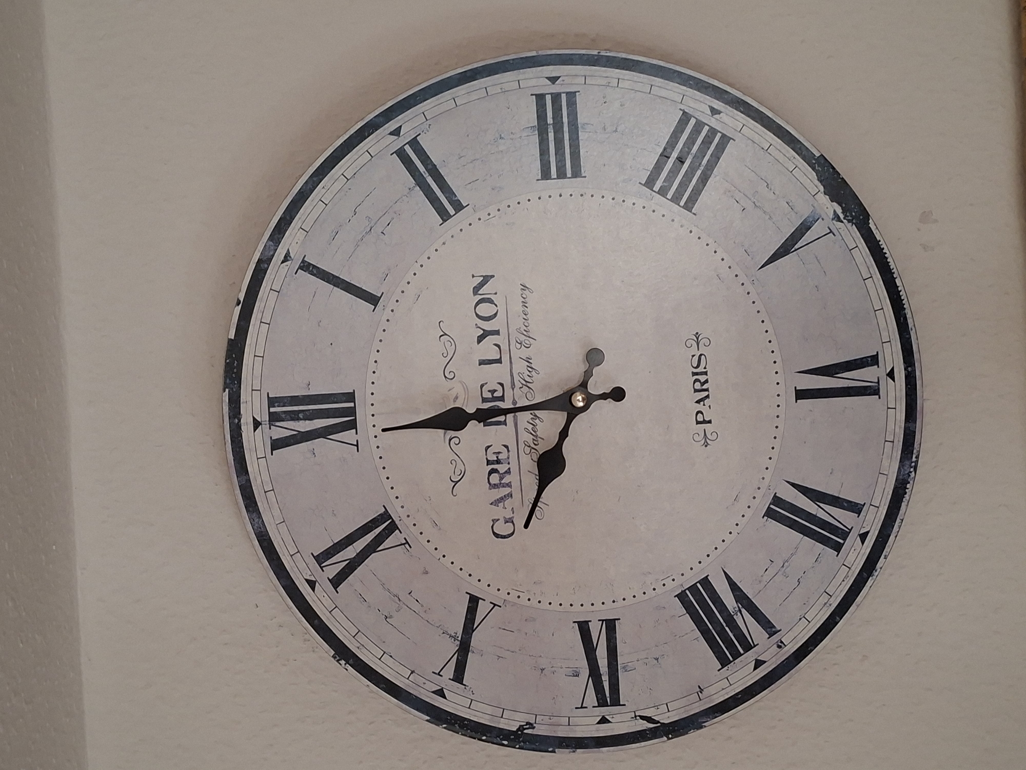 An old, round, wall-mounted clock is displayed against a neutral-colored wall. The clock face is white with black Roman numerals and has the text "GARE DE LYON" and "Speed Safety High Efficiency" printed on it. The clock hands are black; the hour hand points to approximately the 9 o'clock position, and the minute hand points to approximately the 10 o'clock position. The clock has a distressed, antique appearance with visible wear on the edges. The lighting is consistent and appears to be ambient, with no harsh shadows.