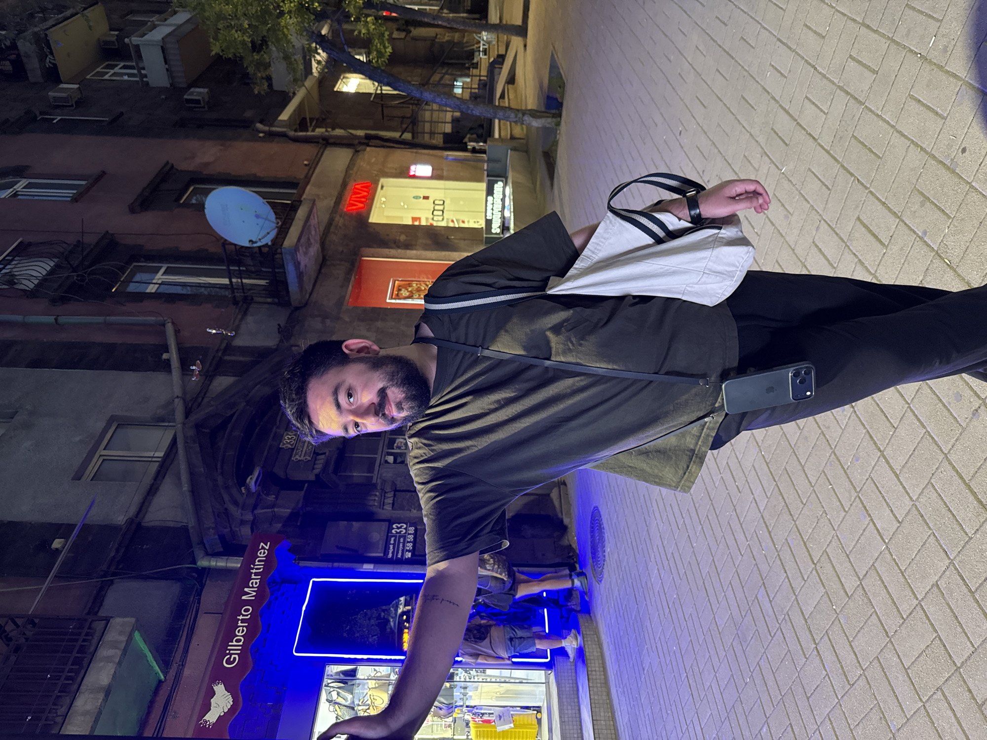 An evening urban scene on Abovyan Street in Yerevan, Armenia, features a male subject posing on a paved sidewalk. The man, with a dark beard and dark hair, wears a dark olive-green t-shirt and dark pants. An iPhone 15 Pro Max, secured by a black lanyard phone case, hangs at his right hip, and a light-beige canvas tote bag with black and white striped handles is slung over his right shoulder. A black smartwatch is visible on his right wrist. He faces the camera with a slight smile, his left arm extended to the left, resting against a dark vertical surface adjacent to a store entrance.
The background immediately behind the subject shows a brightly illuminated store entrance outlined by a vivid blue neon light. Above this, a red awning displays "Gilberto Martínez" in white text and a white handshake logo. To the right of the entrance, affixed to the building, a sign indicates the address "33 Abovyan str." in both Armenian and Latin scripts, along with the phone numbers "58 58 88". Inside the store, two indistinct figures are visible near displays of what appear to be bags or accessories. The sidewalk is paved with light-colored rectangular pavers arranged in a herringbone pattern, with a circular metal manhole cover visible to the left of the man's feet.
Buildings line the street; the immediate building on the left features a reddish-brown facade with darker upper sections. Further down the street on the right, another multi-story building displays a blue satellite dish mounted on a balcony and a red "VIVA" sign at street level. Trees with dark trunks and some green foliage are visible on the far right side of the street, further back. The scene is illuminated by various artificial lights, indicative of nighttime in a commercial urban area.