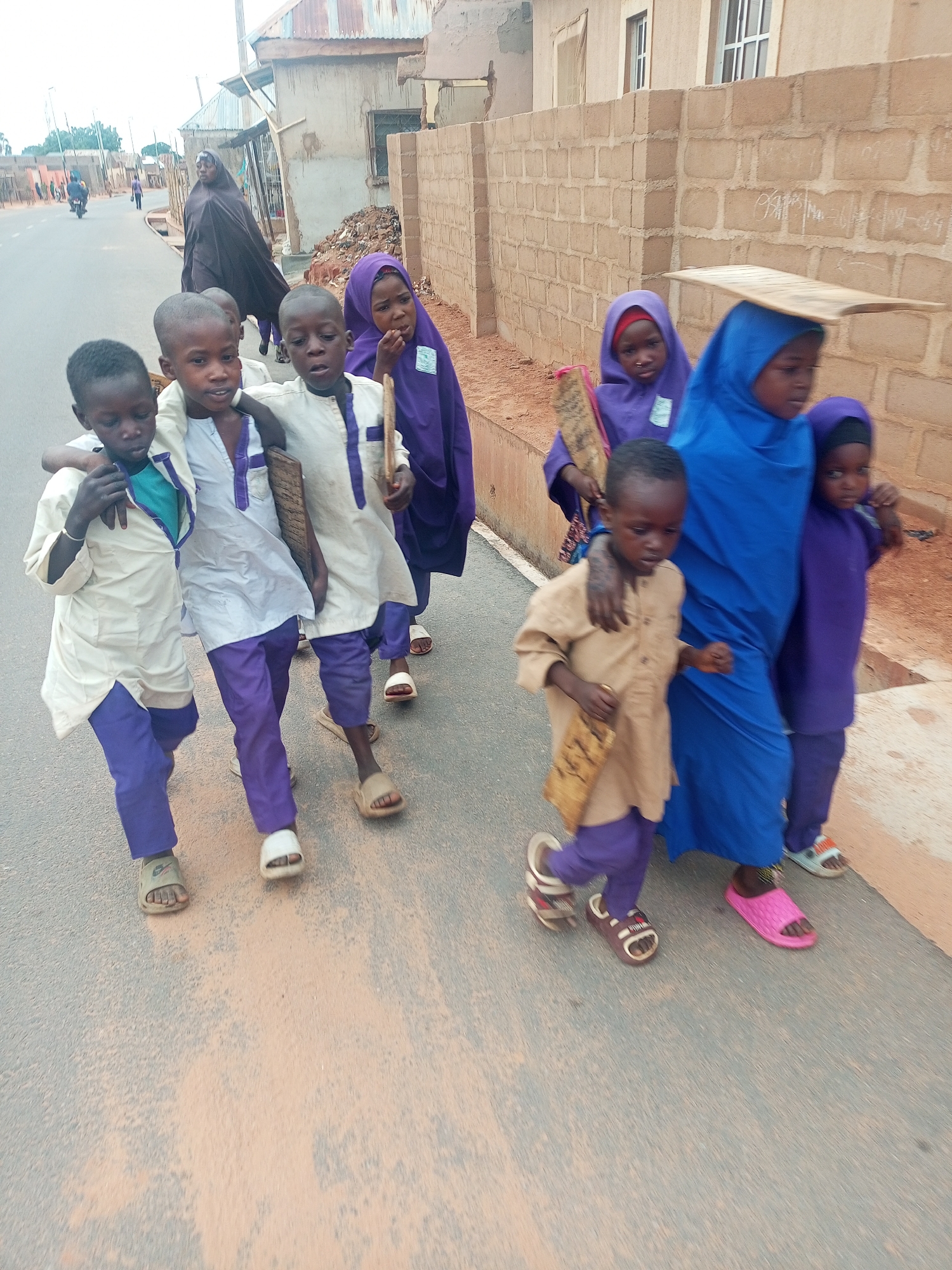 A group of children walk along a street in Tempure, Nigeria. Three boys walk abreast, the boy on the left wearing a white shirt with purple trim, and purple pants. His two friends are dressed similarly but wear white shirts. They have their arms around each other's shoulders. A girl wearing a purple hijab walks behind them, and to the right is a group of children including two more girls wearing hijabs, one purple and one blue, and a young boy wearing a tan shirt with purple pants. The girl in the blue hijab is carrying a flat, rectangular object on her head. In the background, a woman in a black robe walks in the same direction as the children. The road is paved and the surrounding buildings appear to be residential.