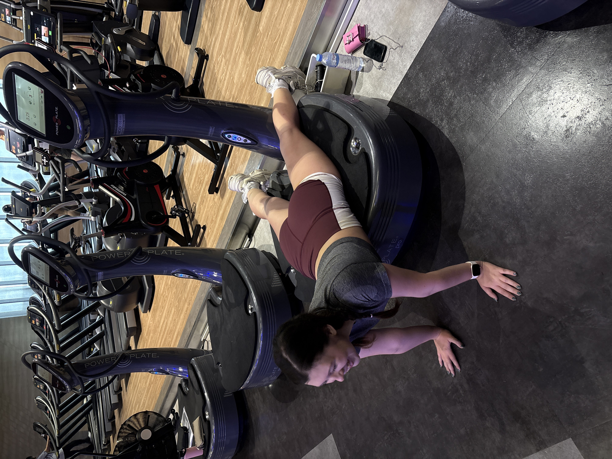 A woman is performing an exercise on a Power Plate machine in a gym. She is in a prone position, with her hands on the floor in front of her and her feet resting on the vibration plate. She is wearing a grey t-shirt, maroon shorts, and white athletic shoes. She has dark hair pulled back in a braid and is smiling. In the background, other Power Plate machines and treadmills are visible. The floor is dark grey, and the walls are light-colored with large windows. The lighting is bright, suggesting it is daytime. The gym appears to be modern and well-equipped. The overall scene depicts a fitness routine in progress.