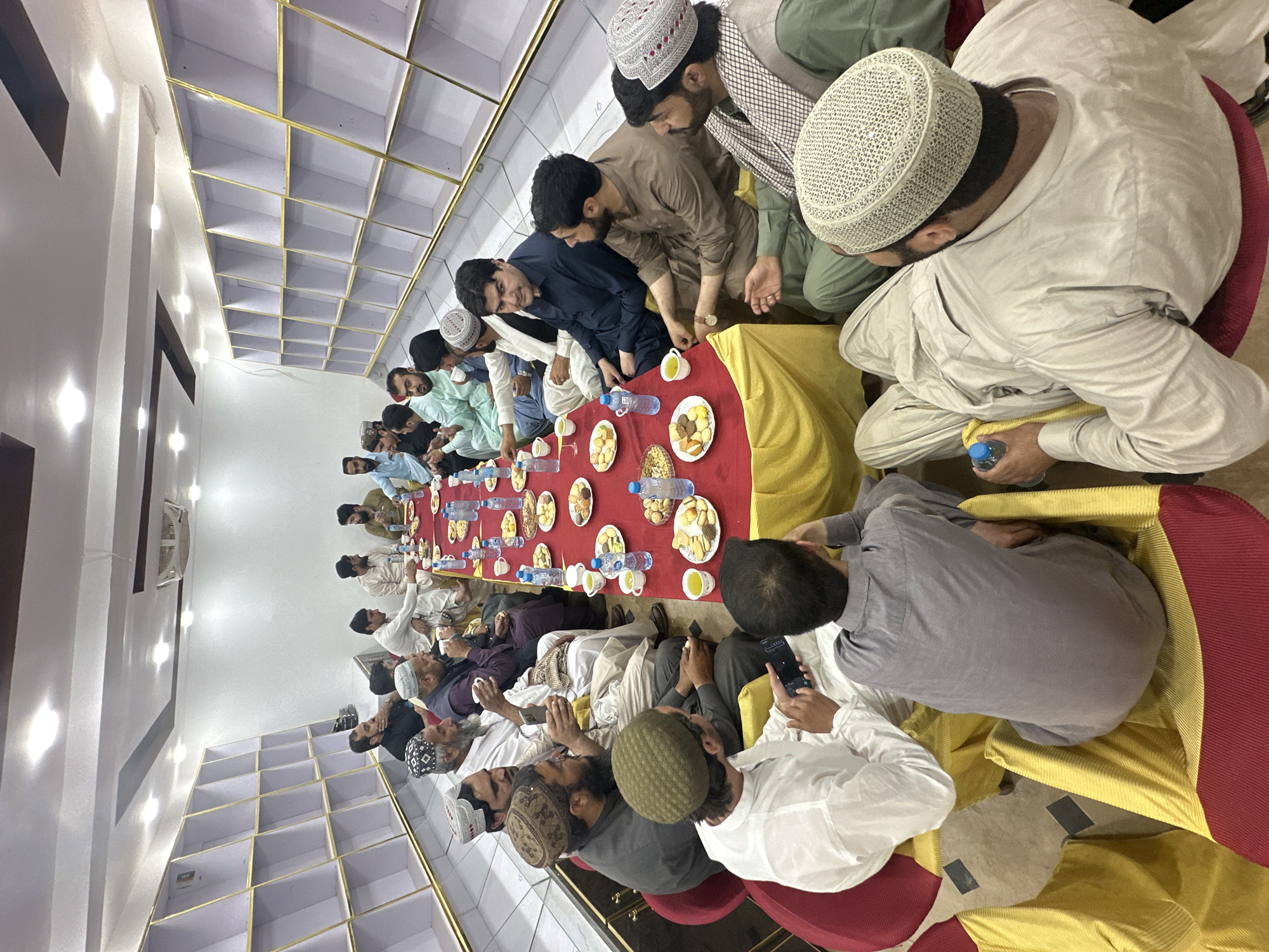 A group of approximately 25 men are seated on the floor around a long, red-covered table laden with food and water bottles, inside a room in Quetta City Tehsil, Pakistan. The men are dressed in various traditional Pakistani clothing, including kurtas and head coverings, sitting on cushions of red and gold fabric. Some of the men are holding or looking at their phones. The room has a white ceiling with a grid-like pattern.