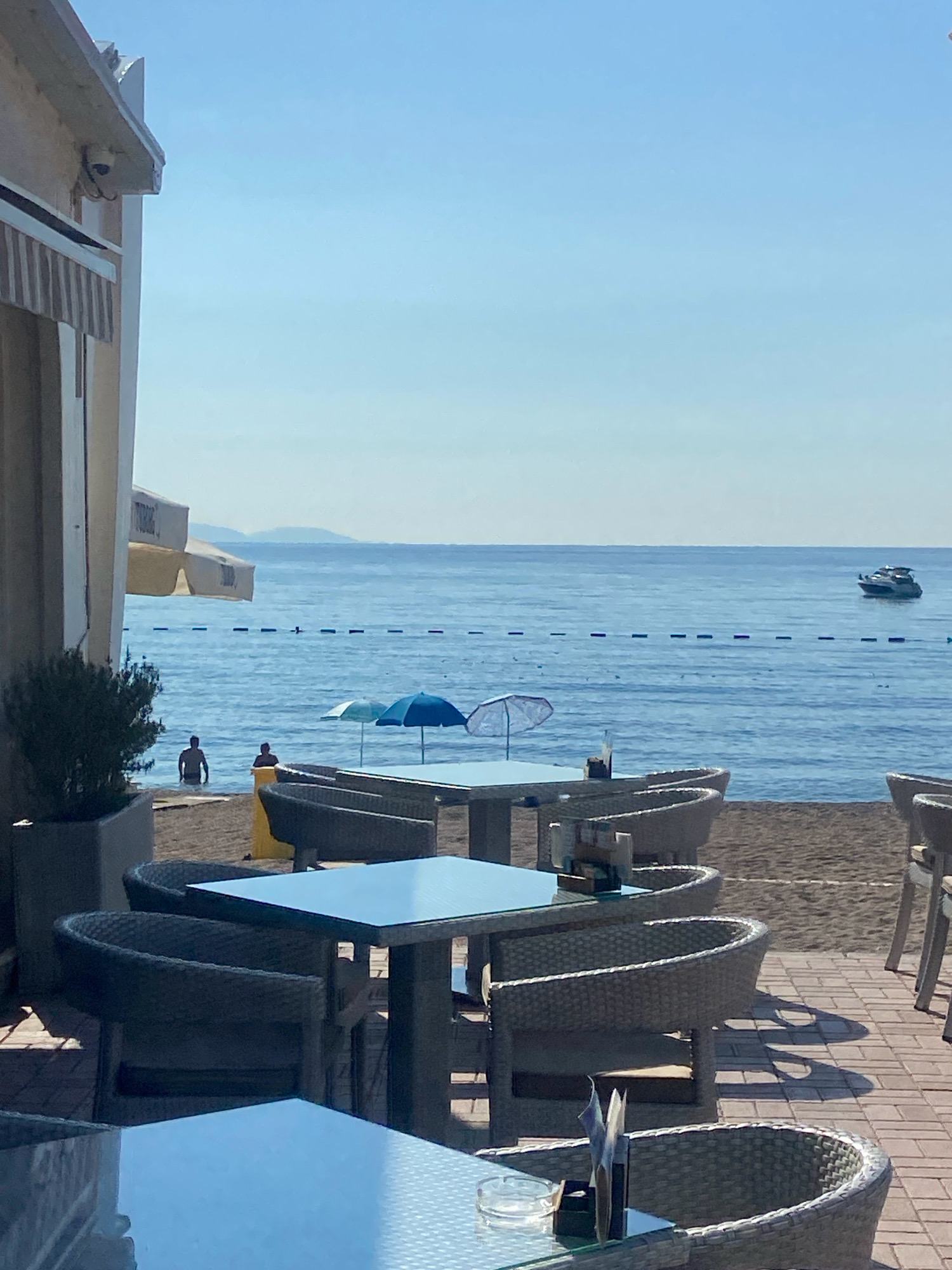This is a photograph taken from a seated perspective, looking out towards a beach and the sea under a clear blue sky. The foreground features outdoor seating at a restaurant or cafe, with multiple tables and chairs. The tables have glass tops, and some have menus or ashtrays on them. The chairs are made of wicker-like material. To the left, a large potted plant is visible.
Beyond the seating area is a sandy beach. In the water, two individuals are present, standing partially submerged. Several beach umbrellas are set up on the sand, one light blue and one darker blue. Further out in the sea, a white motorboat is visible. A line of buoys or markers stretches across the water. In the distance, a faint outline of hills or mountains can be seen against the horizon. The overall impression is of a sunny day at a seaside establishment. The photograph appears to have been taken in Prijevor, Montenegro.