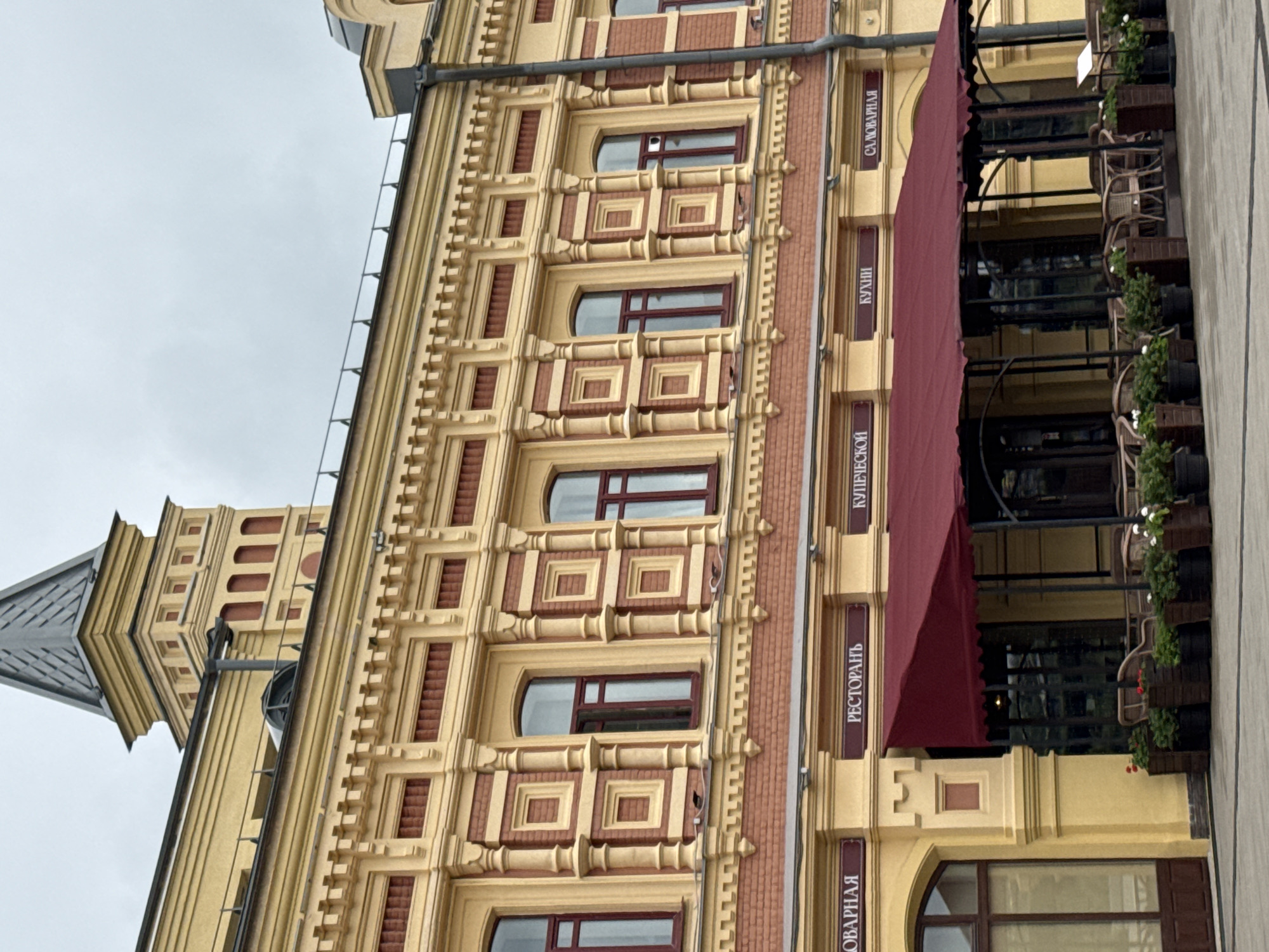 The image shows the facade of a yellow building with ornate architectural details in Nizhny Novgorod, Russia. The building has multiple stories with arched windows framed by columns and decorative brickwork. Above the first-floor windows, there are signs in Russian that read "ТОВАРНАЯ" (Grocery), "РЕСТОРАНЪ" (Restaurant), "КУПЕЧЕСКОЙ" (Merchant's), "КУХНИ" (Kitchen), and "САМОВАРНАЯ" (Samovar Room). A deep red awning extends over the entrance to what appears to be a restaurant or cafe, with outdoor seating visible beneath it. Planters with green foliage and white flowers are placed in front of the seating area. The sky is overcast, suggesting a cloudy day.