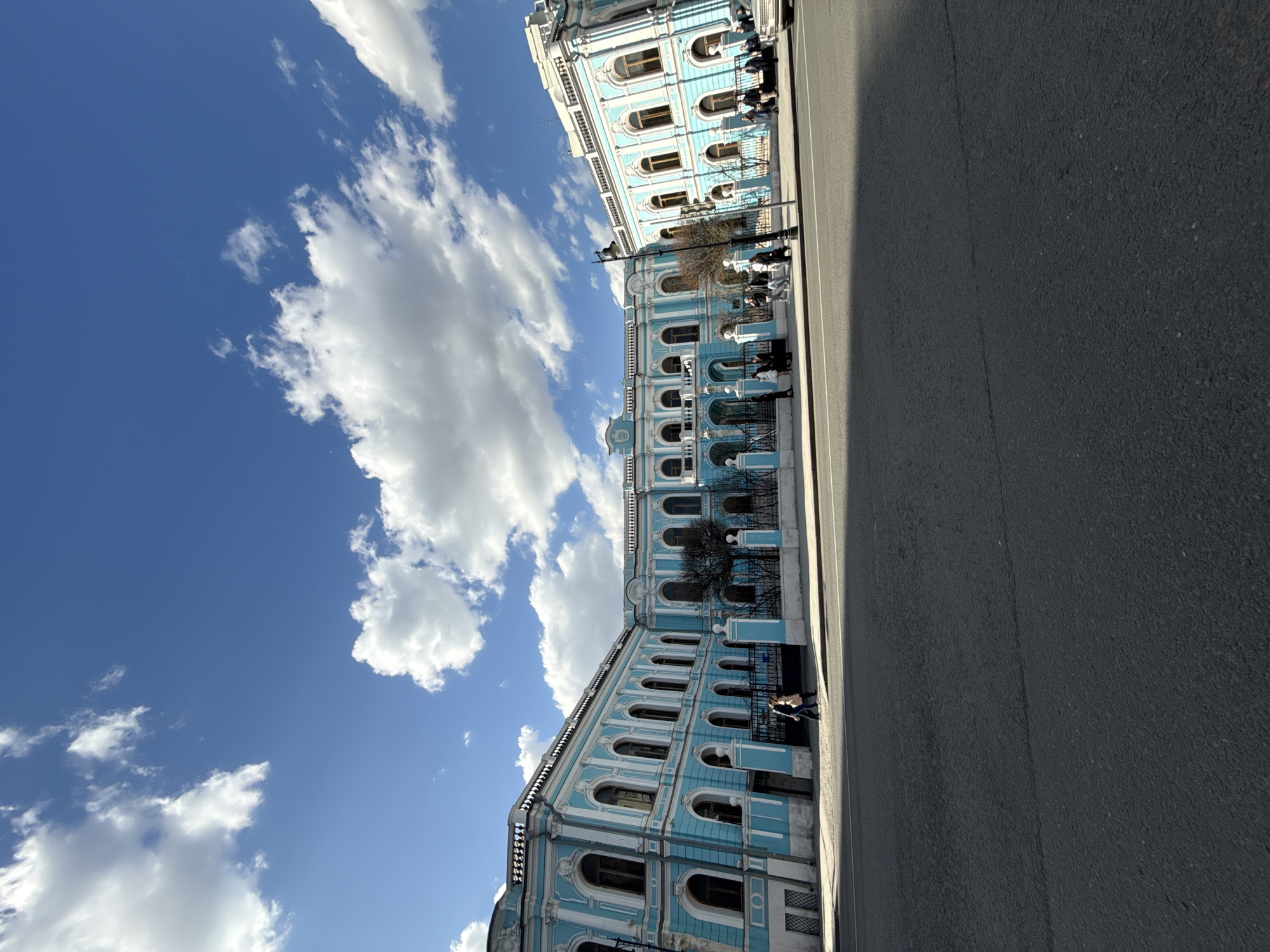The image shows a building in Moscow, Russia, specifically the Smolny Institute. The building has a light blue facade with white trim and is set against a bright blue sky with fluffy white clouds. A paved road stretches along the right side of the image. People are sitting on benches on the building's terrace.