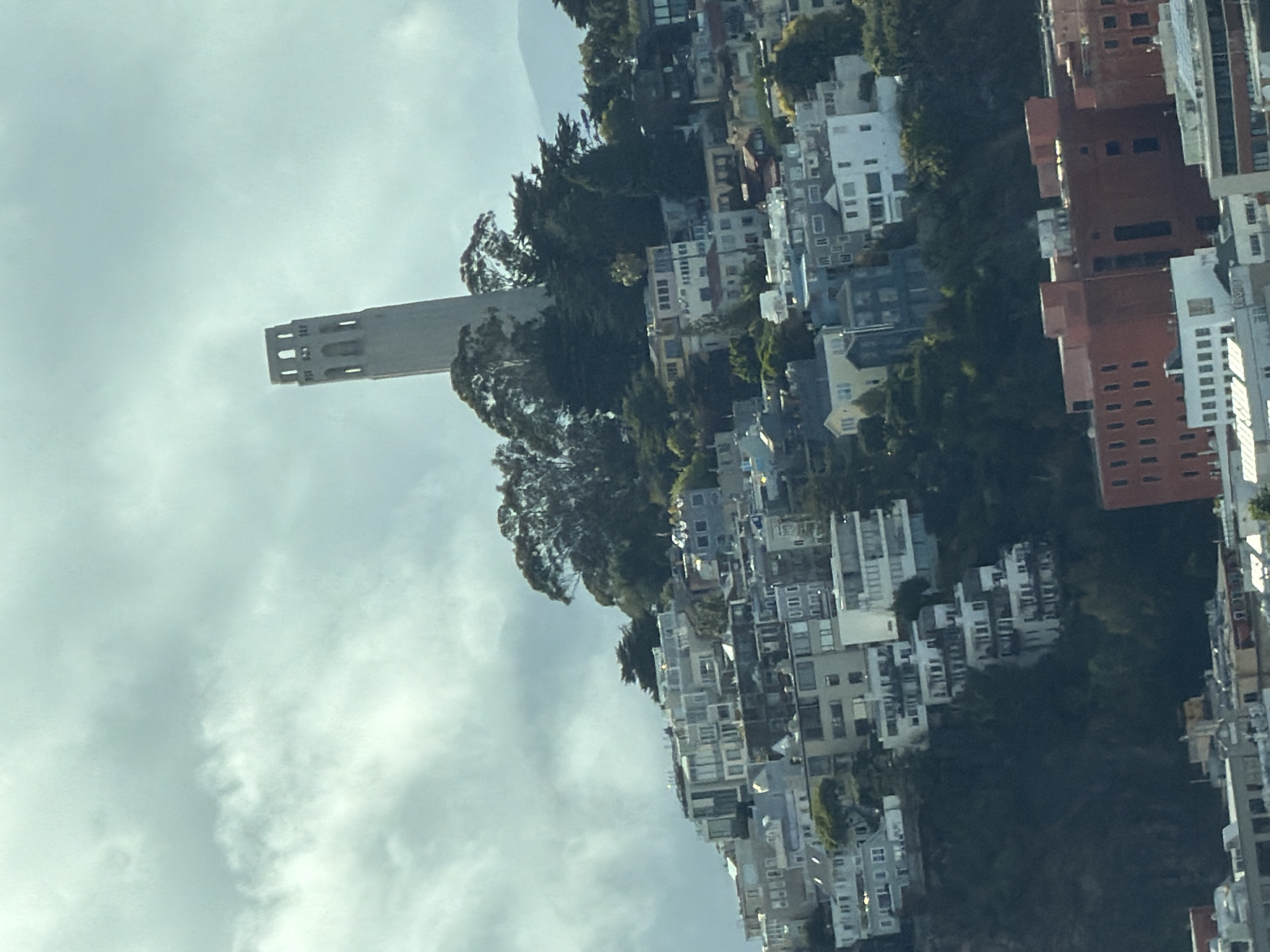The image shows a panoramic view of the city of San Francisco, California, USA, with a focus on Telegraph Hill and Coit Tower. The architecture is characteristic of San Francisco's hilly terrain, with densely packed buildings cascading down the slopes.
The most prominent landmark is Coit Tower, a cylindrical concrete tower with an observation deck at the top, situated on Telegraph Hill. The tower is light-colored and stands out against the cloudy sky.
The hillside is covered with numerous buildings of various sizes and styles, indicating a mix of residential and possibly commercial properties. Many of these buildings have white or light-colored facades, with multiple windows and balconies. The dense urban development covers the hillside, creating a textured urban landscape.
Trees and foliage are interspersed among the buildings, particularly on the steeper slopes and around Coit Tower. The vegetation appears lush, suggesting a temperate climate.
In the foreground, at the bottom of the image, there are more buildings, some of which are larger and appear to be more modern or industrial, characterized by red brick facades and a more geometric design.
The sky is overcast with diffused sunlight, creating a soft, ambient light that bathes the scene. The clouds are textured and varied, with some areas of brighter white and others of darker gray, suggesting a dynamic weather system. The overall impression is one of a sprawling urban environment built on challenging terrain, with a significant landmark punctuating the skyline.