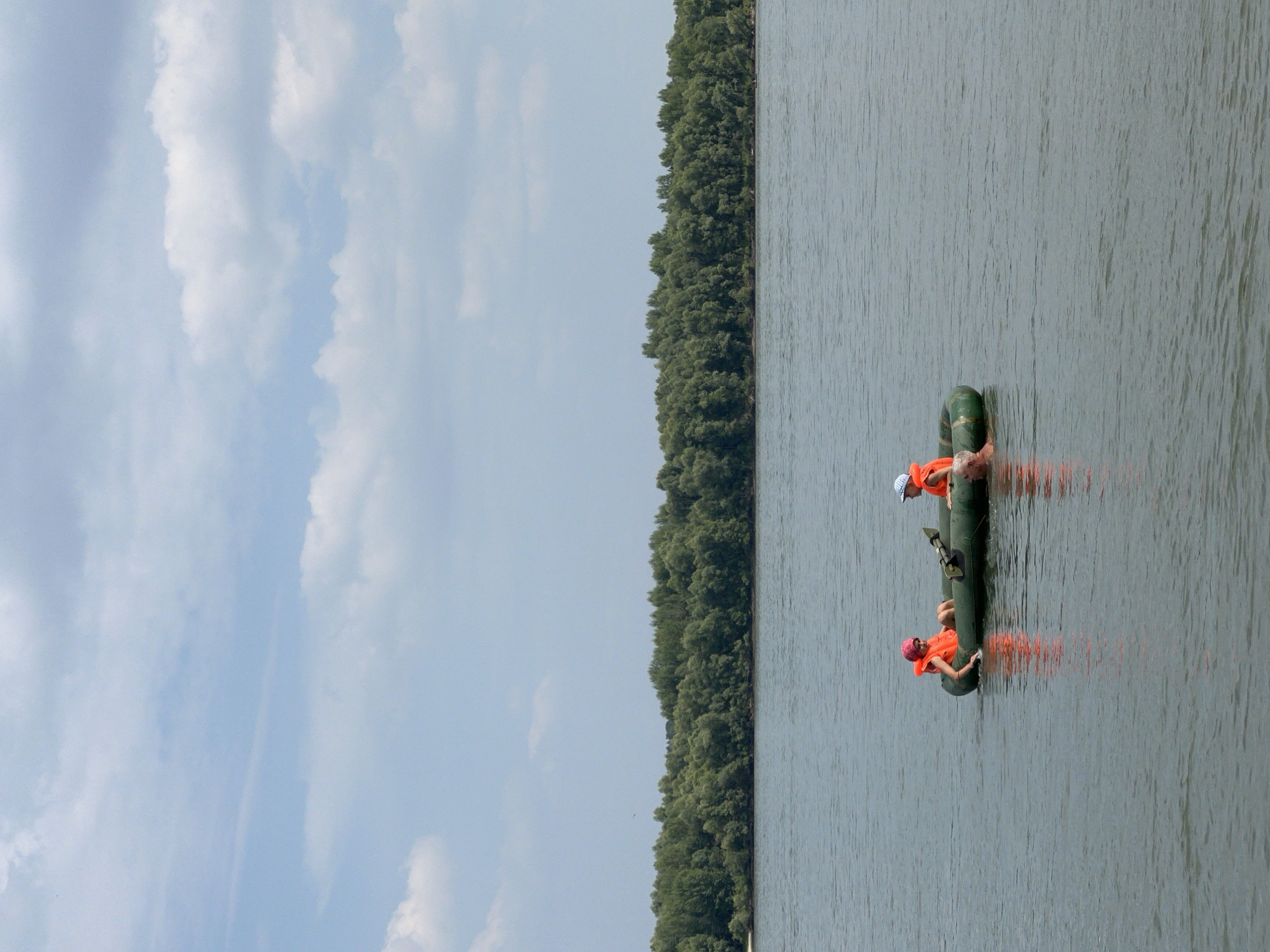 The image shows an inflatable raft with three people on a body of water. The raft is green, and two of the individuals are wearing orange life vests. One person is seated in the front left of the raft, wearing a pink head covering and an orange life vest. Another person is seated in the front right, wearing a white cap and an orange life vest. A third person's head and shoulders are visible in the water to the right of the raft. The water is calm and gray, reflecting the overcast sky which is filled with white clouds. In the background, a dense line of green trees forms a treeline along the far shore. The overall scene depicts an outdoor recreational activity on a river or lake, possibly near the city of Mochishche, Russia, given the treeline and the nature of the water.