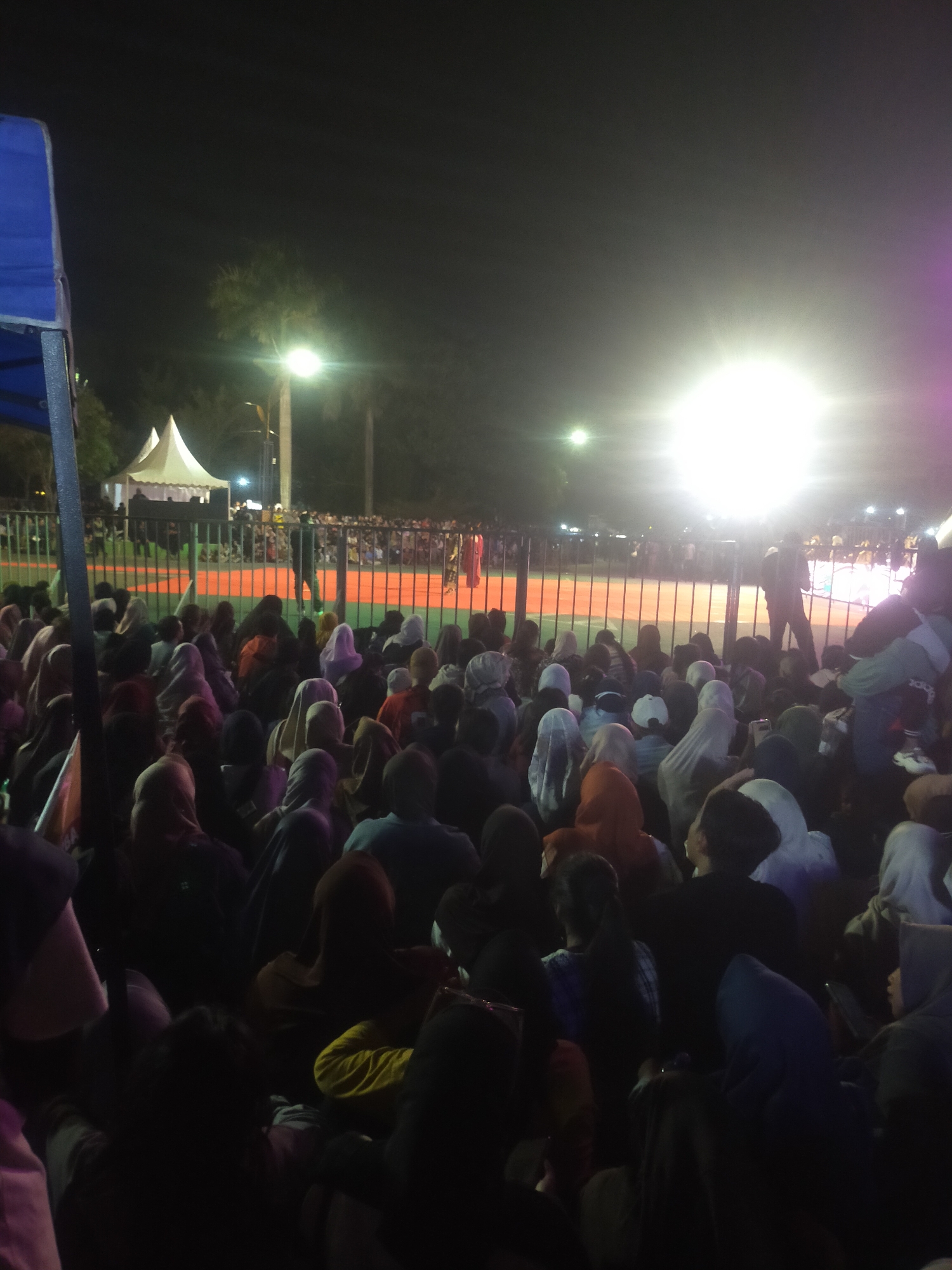 The image depicts a night scene, likely an outdoor event, with a large crowd of people seated and standing. The foreground is filled with people, many wearing headscarves, indicating a predominantly Muslim gathering. The crowd is looking towards a brightly lit area in the background, suggesting a stage or a focal point of the event.
In the midground, there's a fence separating the crowd from an area with a red surface, possibly a sports court or performance space. Beyond the fence, several white tents are visible, and there are more people gathered around them. A bright light source illuminates the area, creating a glow and lens flare. The background shows trees and the night sky.
The overall impression is of a community gathering or event taking place after dark. The presence of tents and a bright light suggests it could be a religious ceremony, a festival, or a public lecture. The crowd appears engaged and attentive to the illuminated area.
Given the prompt's request for information about the location in the city of Slawi, Indonesia, and without explicit visual cues in the image itself to confirm this specific location, it's impossible to definitively identify it as Slawi. However, such events are common in various cities in Indonesia. Without further context or visible landmarks, any attribution to Slawi would be speculative.
