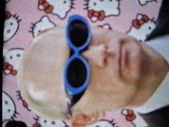 A still image depicts an adult male, identified as resembling Vladimir Putin, positioned from the chest up against a vibrant pink background adorned with a repeating pattern of white Hello Kitty cartoon faces. The subject has light skin, a prominent bald head, and wears a dark suit jacket over a white collared shirt. He is outfitted with distinctive blue-framed, dark-lensed oval sunglasses. His gaze is directed forward, and his facial expression is neutral. The visual quality suggests a photograph taken of a digital screen, exhibiting minor pixelation. The location is Unknown, Unknown.