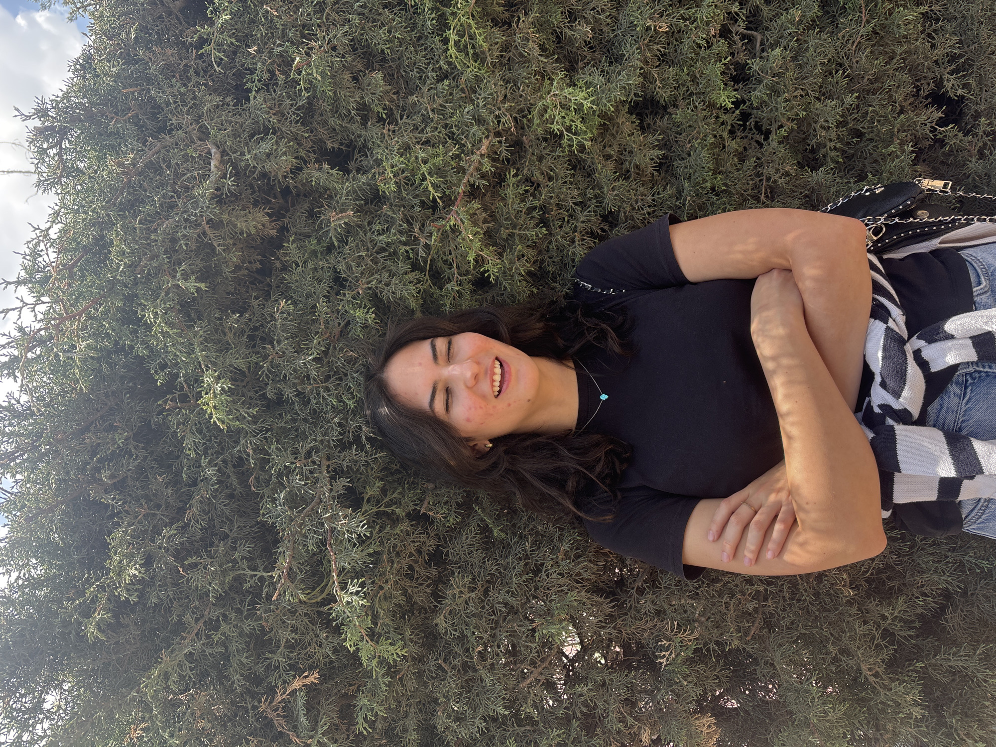 A young woman is lying on her left side in front of a large green bush. The woman has dark curly hair and is wearing a black t-shirt and jeans. She is smiling, with her eyes closed. Her arms are crossed, and a black and white striped sweater is draped over her shoulder, a black purse at her side. The bush is full and lush, and the sky is visible in the top left corner of the image. The location is San Juan Arcos Ojo de Agua, Mexico.