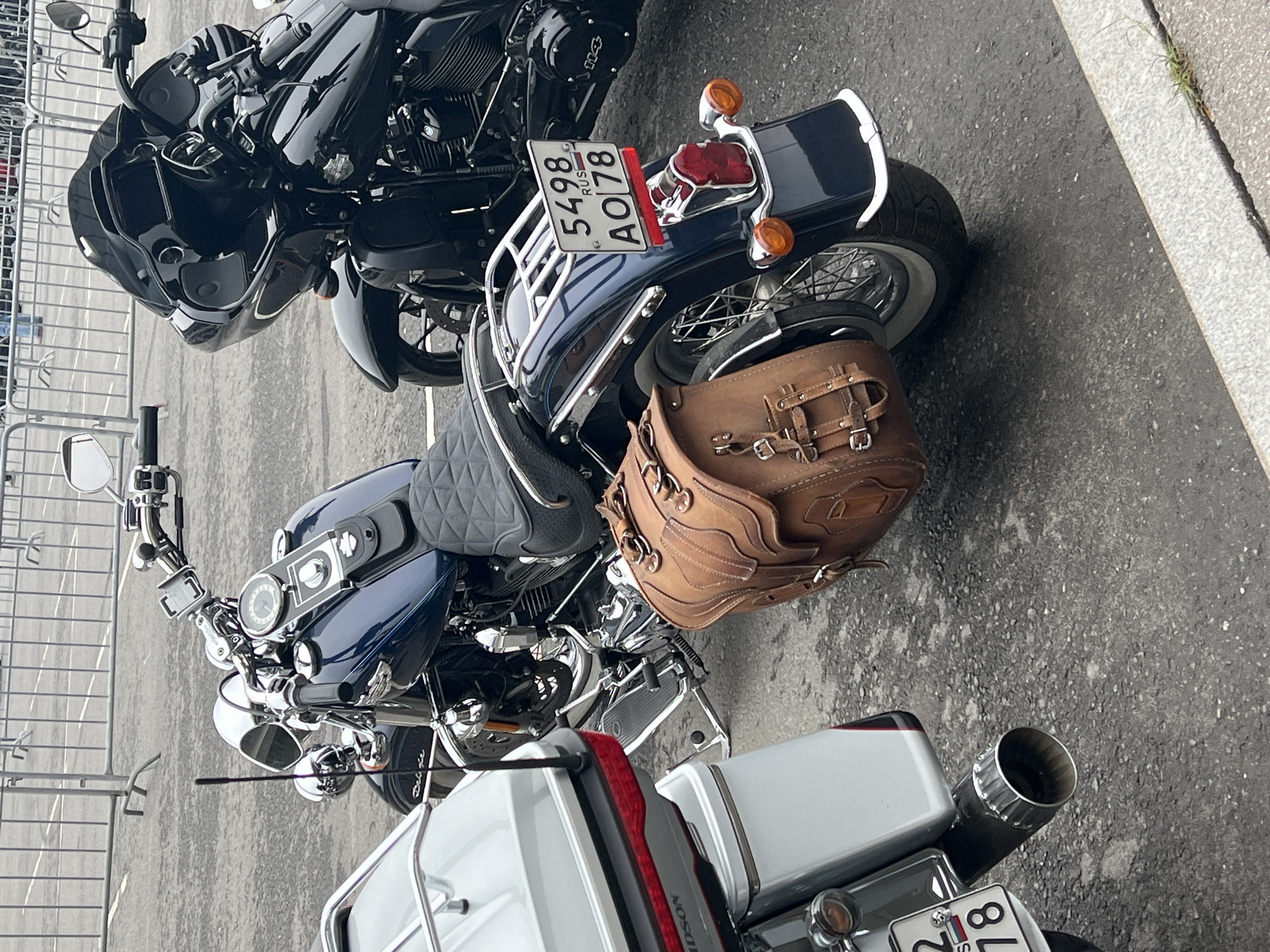 The image shows a parking area with three motorcycles, likely located in Saint Petersburg, Russia. The motorcycles are parked closely together. The license plates on the vehicles suggest they may be registered in Russia. The motorcycles feature a variety of details, including leather saddlebags, chrome accents, and custom modifications. The overall environment appears to be an urban setting, with a metal fence visible in the background.