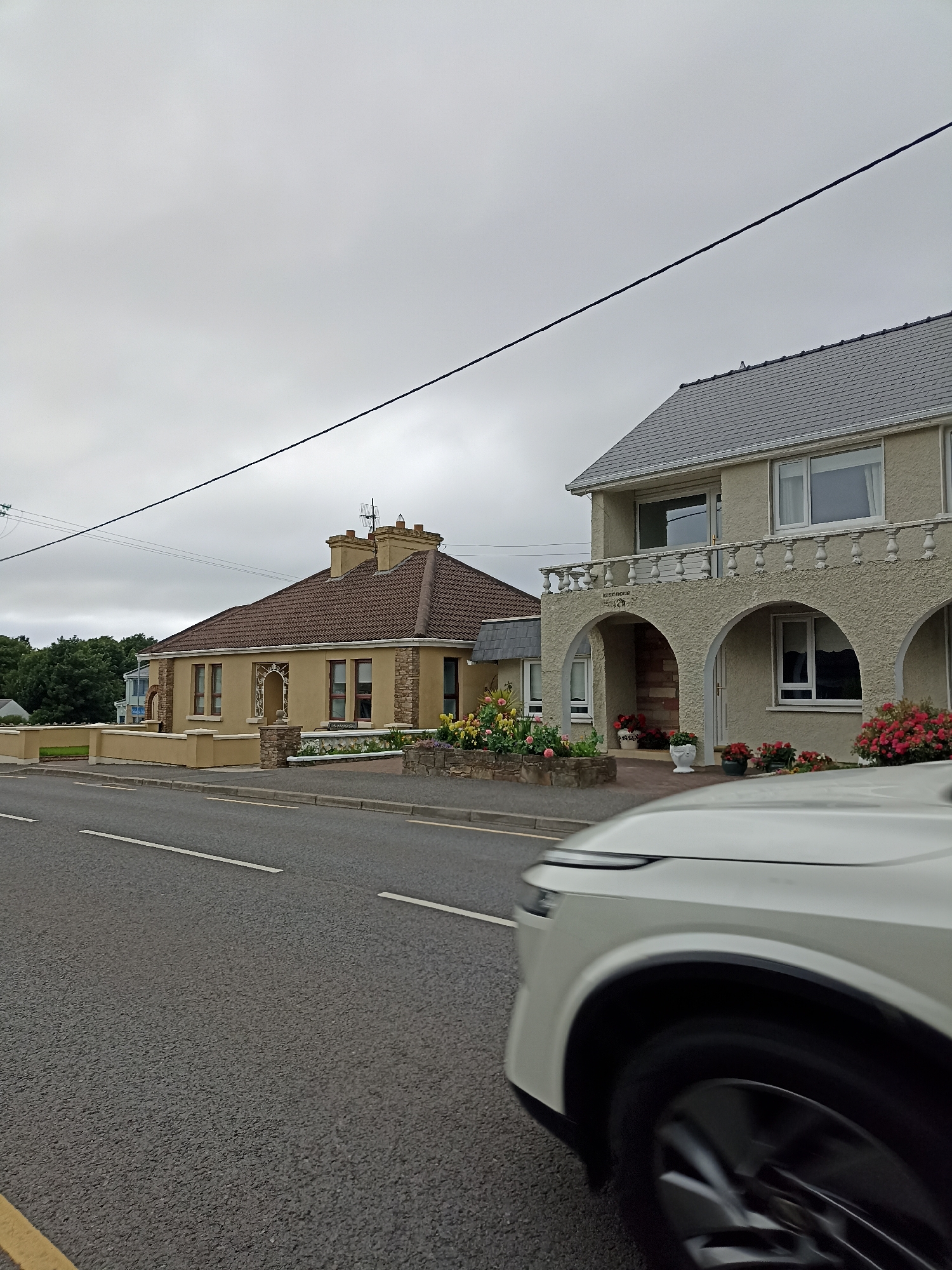 A front-left view captures a white vehicle driving on a paved road in Annagry, Ireland. The road features white lane markings and a yellow line on the left edge. Adjacent to the road are two houses. The house on the left is a single-story structure with a brown, tiled roof, light yellow walls, and stone accents. It has several windows and a low, beige-colored wall in front. The house on the right is a two-story building with a grey, tiled roof, a stucco exterior, and arched openings on the ground floor. A balcony with a white railing runs across the second story. Both houses have flower beds with red and yellow blooms. The sky is overcast and grey, and a power line runs diagonally across the upper portion of the image. Trees are visible in the background.