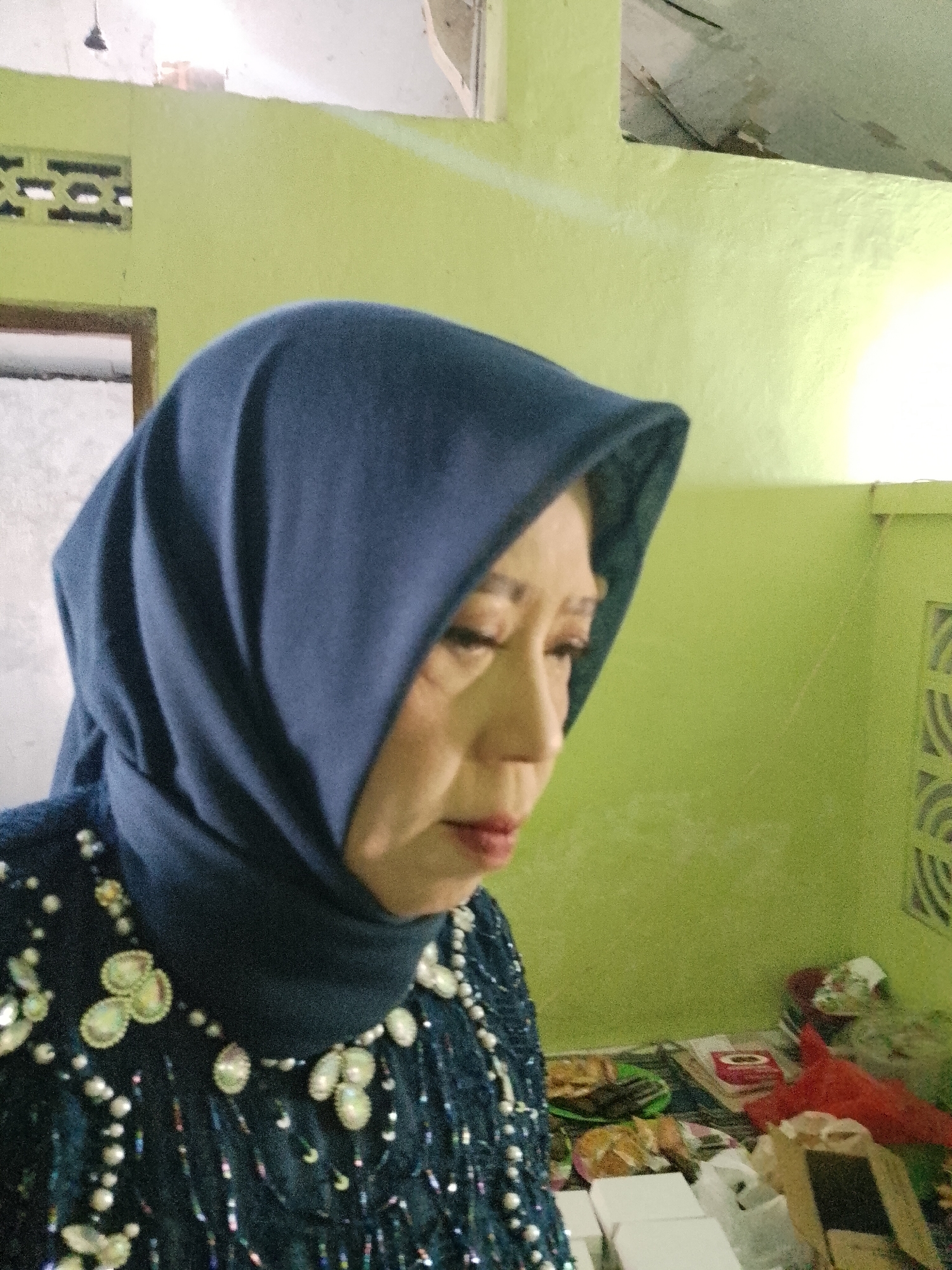 A medium shot captures a woman in a dark blue hijab and an ornate, dark blue dress adorned with pearls and iridescent embellishments. She is looking downwards and to her right, with her mouth slightly open, and her eyes are partially closed, suggesting contemplation or fatigue. Her face displays signs of age.
The background is a light green wall, textured and slightly uneven. To the left, a portion of a window with a wooden frame is visible, with a decorative grille on the wall above it. To the right, the wall continues, and a decorative grille is also present.
In the foreground, on a surface below the woman, are several items that appear to be stacked boxes, some white and some light brown. A red plastic bag, some papers, and other assorted objects are also present, suggesting a domestic or storage setting. The overall lighting is somewhat dim, with a bright light source illuminating the upper right portion of the scene, casting a glow on the wall. The setting appears to be a room in Kecamatan Pati, Indonesia, possibly a home or a small shop.