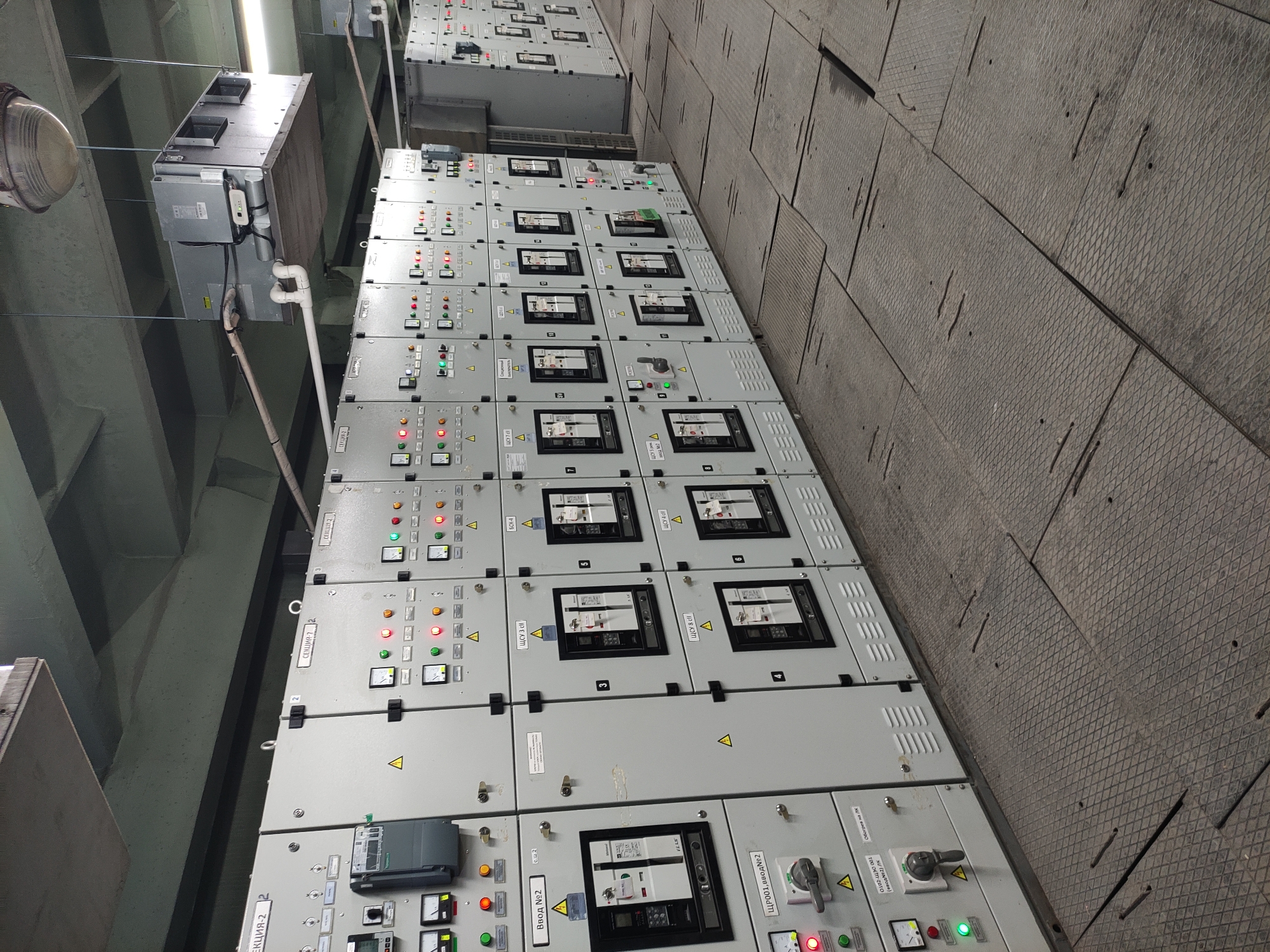 The image shows a series of electrical control panels inside a building, most likely an industrial or utility facility. The panels are light gray and feature various gauges, meters, switches, indicator lights, and digital displays. Warning symbols are visible on several panels. Based on the Russian text written on several panels, the location could be the city of Pavlodar, Kazakhstan. The panels are installed on a gray wall, and there are some overhead fixtures. The panels are aligned in one single large row.