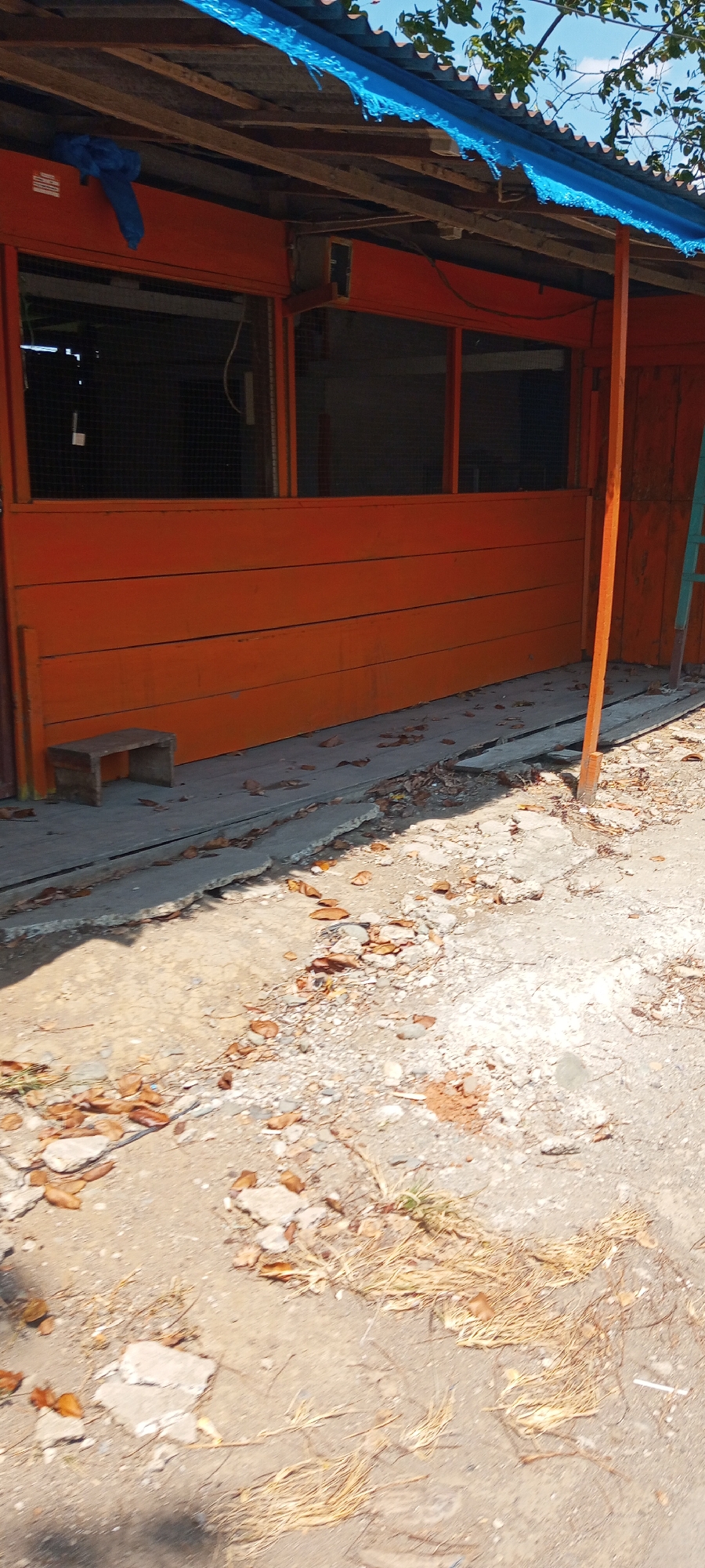 The image shows an outdoor scene in Lempa, Indonesia, featuring a dilapidated roadside structure. The structure appears to be a small shop or stall, with an orange-painted wooden front. The roof is covered with corrugated metal and a tattered blue tarp. Behind the orange planks, there are dark openings, possibly windows or doorways, partially covered with mesh. A small, wooden stool is placed near the left side of the structure. The ground in front of the structure is uneven and appears to be a mixture of dirt, gravel, and broken concrete. Fallen leaves and dry grass are scattered across the ground. The overall impression is one of disrepair and neglect.