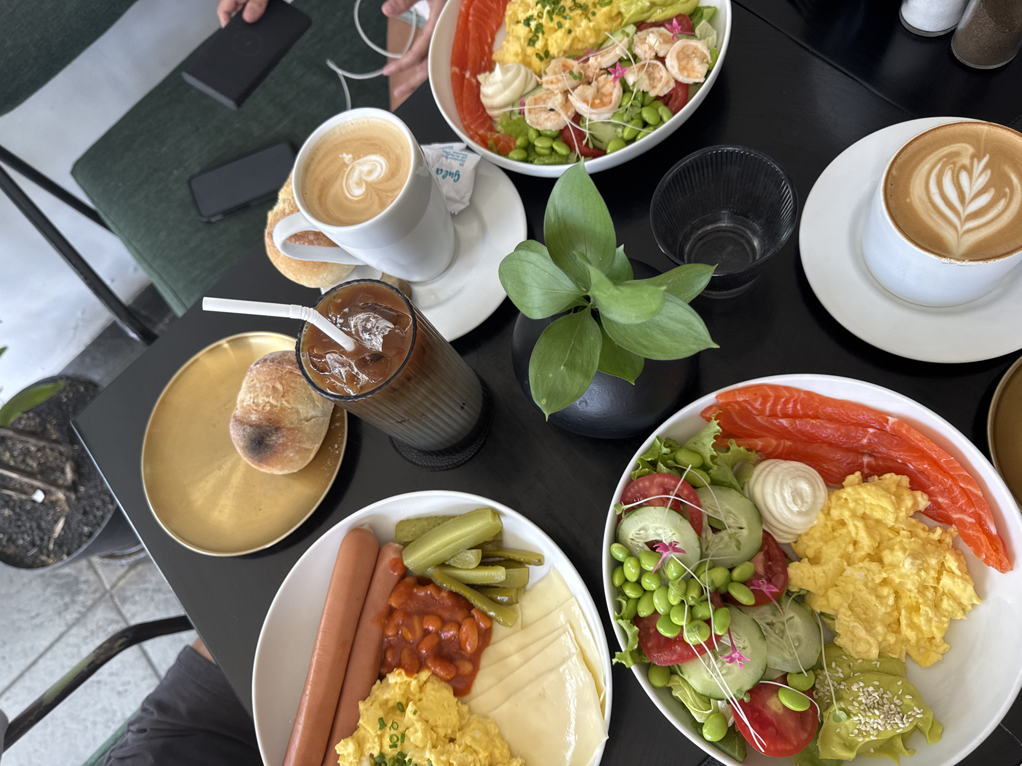 This is an overhead shot of a table laden with an elaborate breakfast or brunch spread, likely at a cafe or restaurant. The dark table surface contrasts with the vibrant colors of the food and white tableware.
In the center, two large white bowls dominate the scene, each filled with a substantial meal. The bowl on the upper side contains scrambled eggs, sliced salmon, shrimp, mixed greens, tomatoes, cucumbers, edamame, and a dollop of mayonnaise or sour cream, garnished with microgreens. The bowl on the lower side features two hotdogs, scrambled eggs, baked beans, sliced yellow cheese, and pickles.
Several beverages are also present. To the left, a white mug holds a latte with intricate heart-shaped latte art, placed on a white saucer with a small white sugar packet visible. Below it, a tall glass of iced coffee with a white straw is positioned on a black coaster. To the right, another white mug with latte art rests on a white saucer. An empty clear glass tumbler is placed near the right latte.
Other items on the table include a gold-colored plate holding a bread roll, a small potted green plant in a black vessel, and a set of salt and pepper shakers in the background. A person's hand is visible in the top left, holding a black phone or power bank, with a charging cable nearby. A dark green upholstered chair and a person's leg are partially visible in the bottom left, suggesting a comfortable seating arrangement.
The scene depicts a relaxed mealtime, with an abundance of food and drinks. The presentation of the dishes, including the latte art and colorful ingredients, suggests an appealing and thoughtfully prepared meal. The only visible text is the brand "Guca" on a sugar packet.