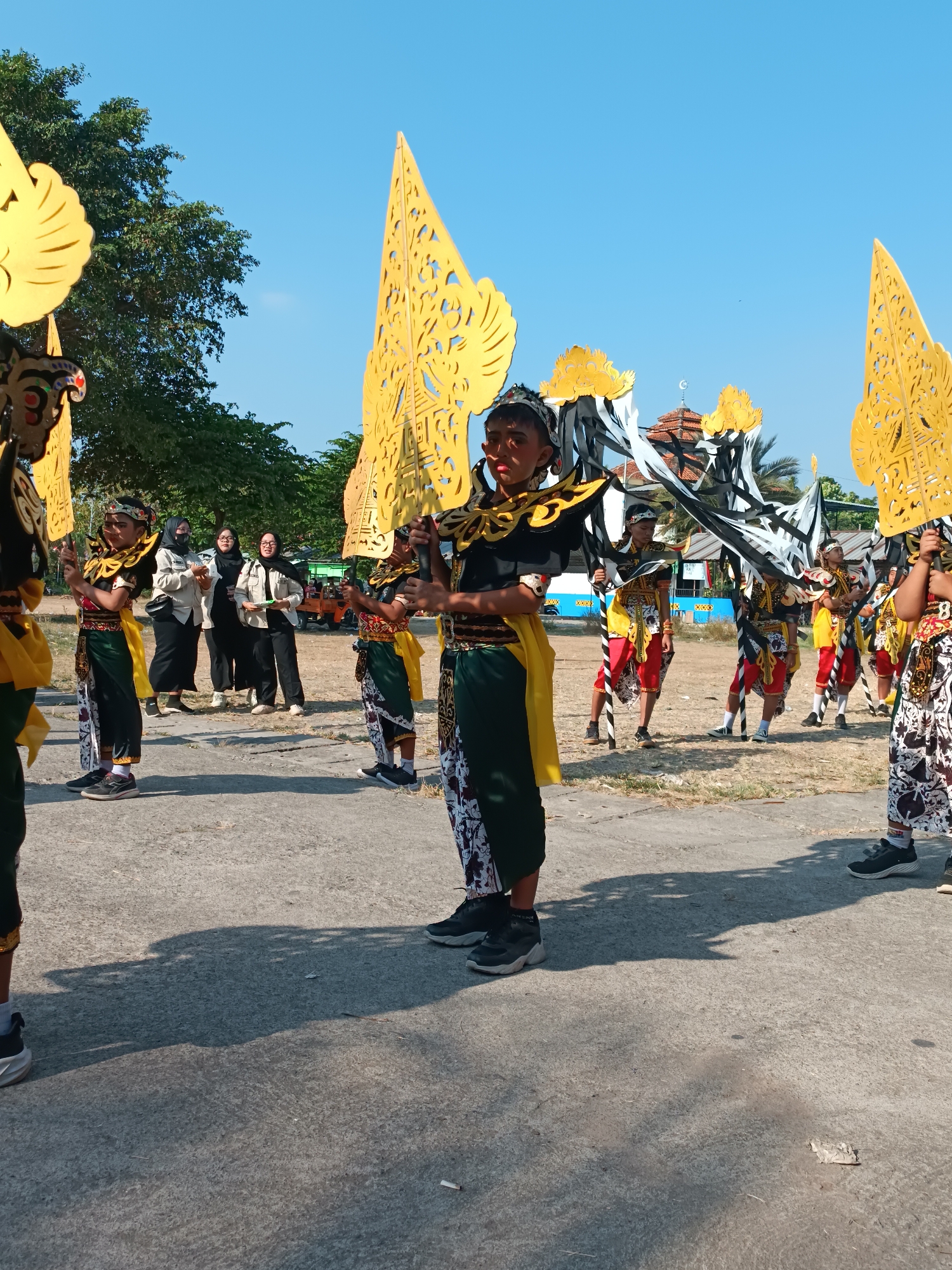 In Imogiri, Indonesia, a group of young people in traditional Javanese attire, resembling Wayang puppets, are participating in an outdoor procession. They are adorned with elaborate headdresses and wear vibrant costumes featuring green, yellow, and red accents, along with patterned pants. Each individual holds a large, ornate, golden fan-like object, characteristic of Javanese cultural performances. In the background, several women in modest clothing, including headscarves, observe the procession. The environment appears to be an open, paved area, possibly a street or courtyard, with trees and a building with a dome and crescent moon symbol visible in the distance, suggesting the presence of a mosque or similar structure. The sky is clear and blue, indicating fair weather. The overall scene depicts a cultural event or celebration taking place in a community setting.