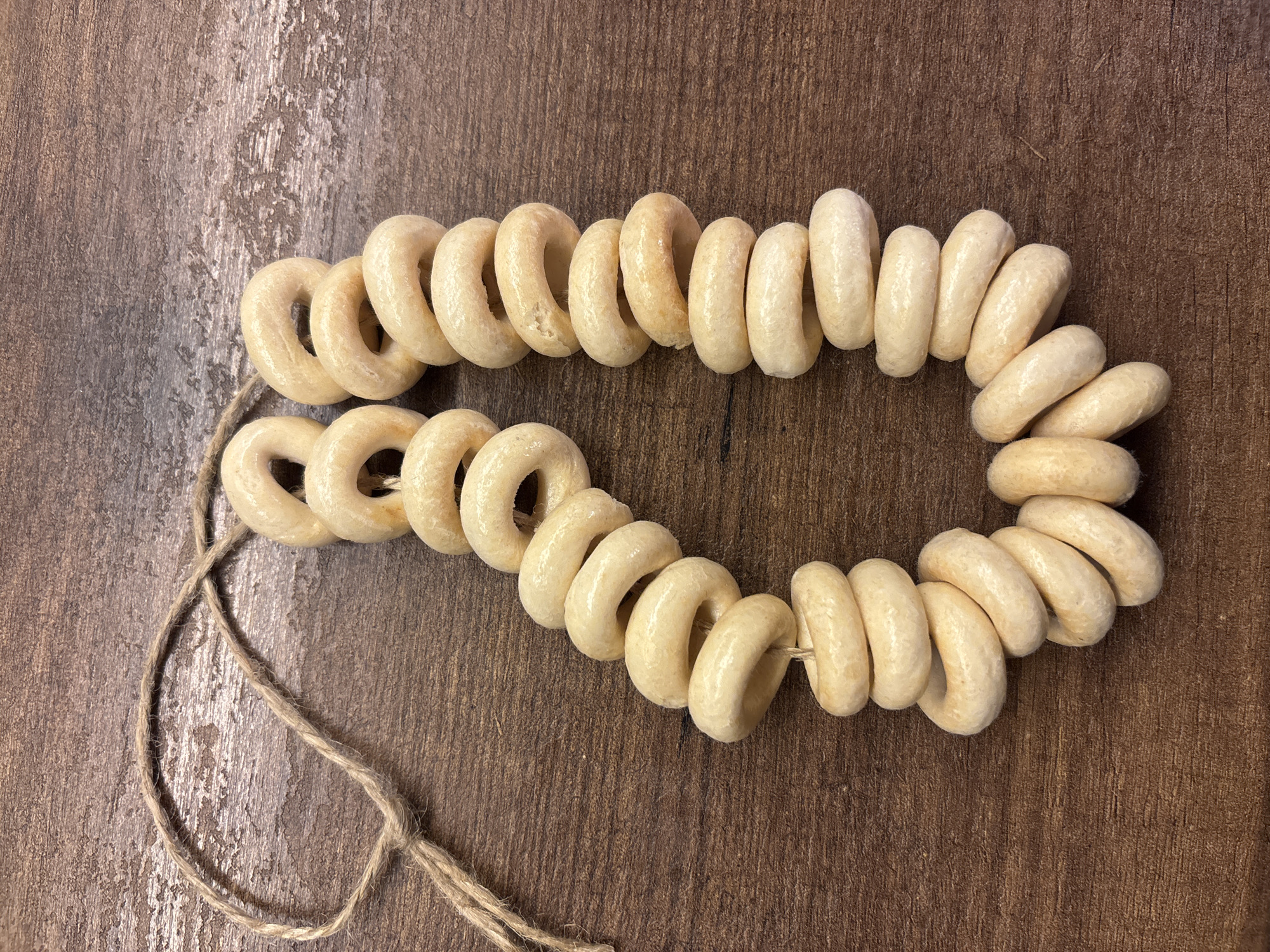 The image presents a still life composition featuring a string of ring-shaped dried biscuits or crackers, commonly known as *sushki* or *baranki* in Russia. These light beige to off-white baked goods are threaded onto a piece of rustic, light-brown twine. The twine forms an elongated, somewhat irregular loop, with the biscuits densely packed along its length.
The entire arrangement rests on a dark brown wooden surface, possibly a table or a board, which displays clear wood grain patterns and variations in tone, suggesting a natural and perhaps aged material. The texture of the wood is visible throughout the background. The lighting is even, casting subtle shadows that highlight the three-dimensional form of the biscuits and the texture of the wooden surface.
No activity or event is actively taking place; it is a static shot of food prepared for consumption or display. The presence of *sushki* strongly cues a cultural context of Eastern Europe, including Saint Petersburg, Russia. There are no people, discernible emotions, or visible text in the image. The scene evokes a simple, traditional, and domestic atmosphere centered around a common Russian snack.