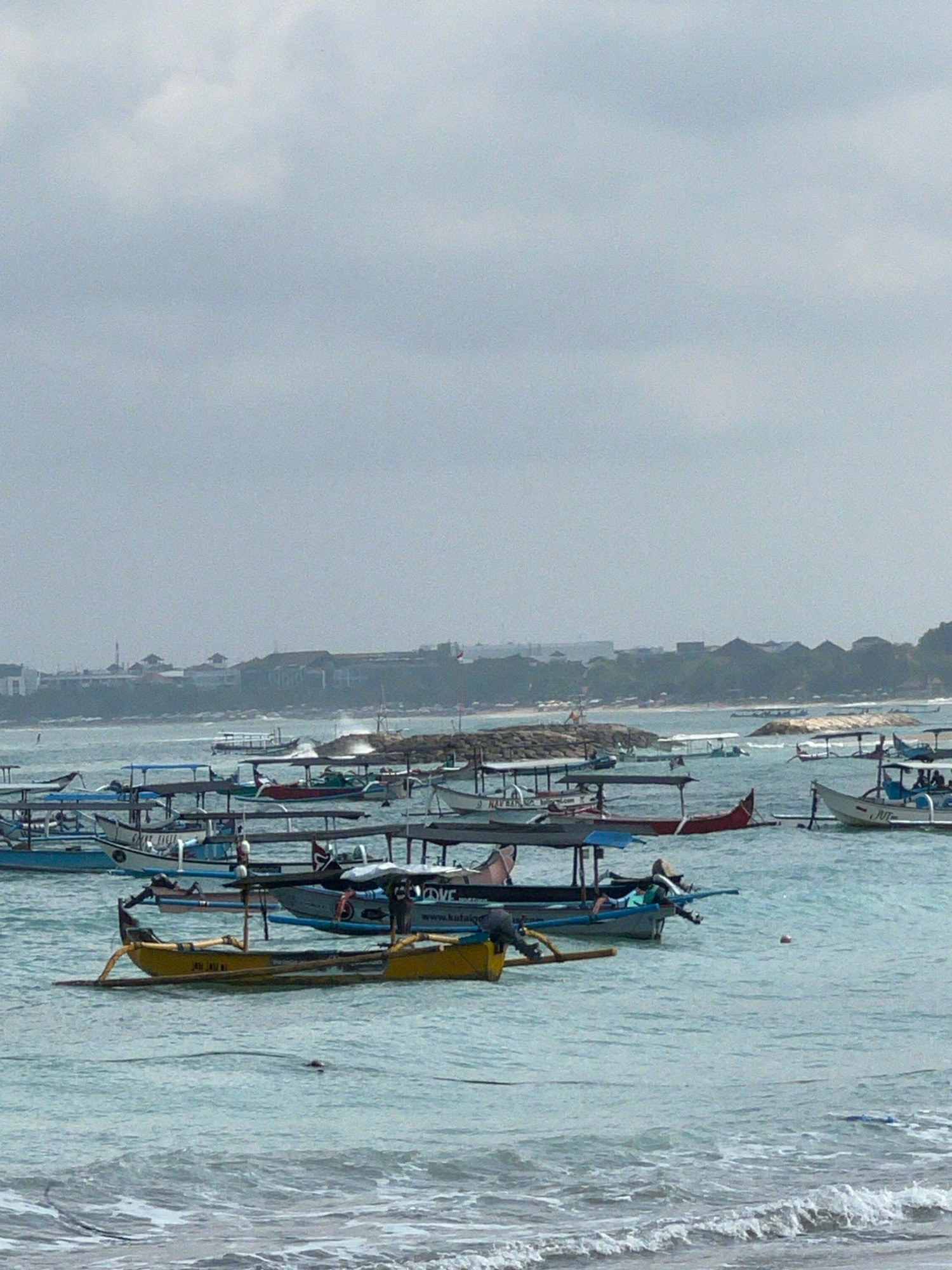 The image shows a harbor full of traditional fishing boats in Kuta, Indonesia. The boats are docked in the calm waters of the sea, with some of them moored together in clusters. The sky is overcast, and the sea is choppy. In the background, a long stretch of coastline is visible, with buildings and trees lining the shore. A rocky breakwater extends into the sea, protecting the harbor from the waves. The boats are of various sizes and colors, with some of them painted in bright hues. They are all equipped with outriggers, which are used to stabilize them in the water. The overall scene is one of a busy fishing harbor, with boats preparing to go out to sea or returning from a fishing trip.