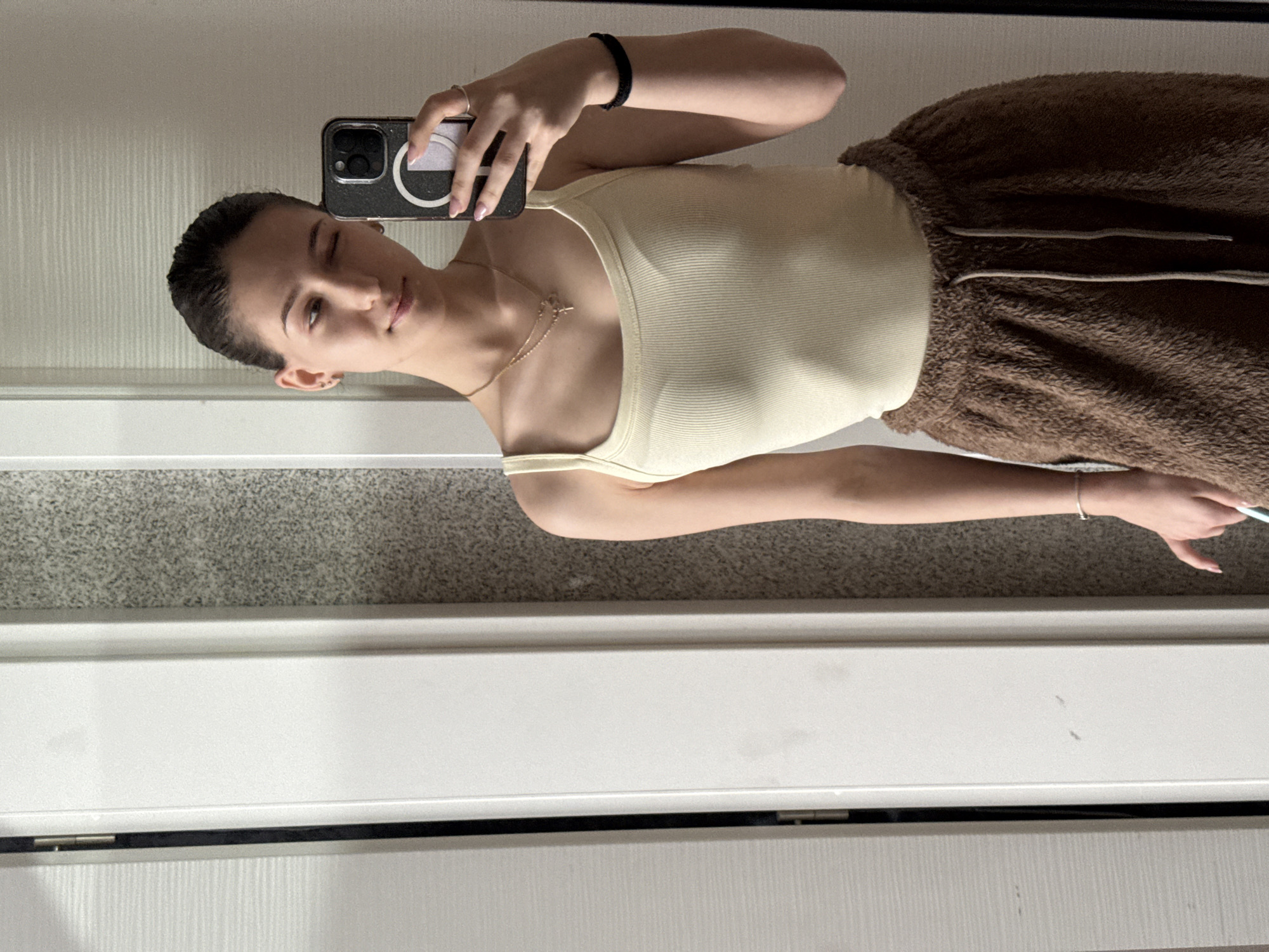 A young woman is taking a selfie in front of a mirror. She is wearing a light yellow tank top and brown fuzzy pants with drawstrings. She holds a black iPhone in her left hand. She is also wearing a black bracelet and a ring. A silver necklace is visible. The environment is plain, with a white wall and a gray, textured surface beneath. The location is not discernable from the image and cannot be determined to be in Sofia, Bulgaria.