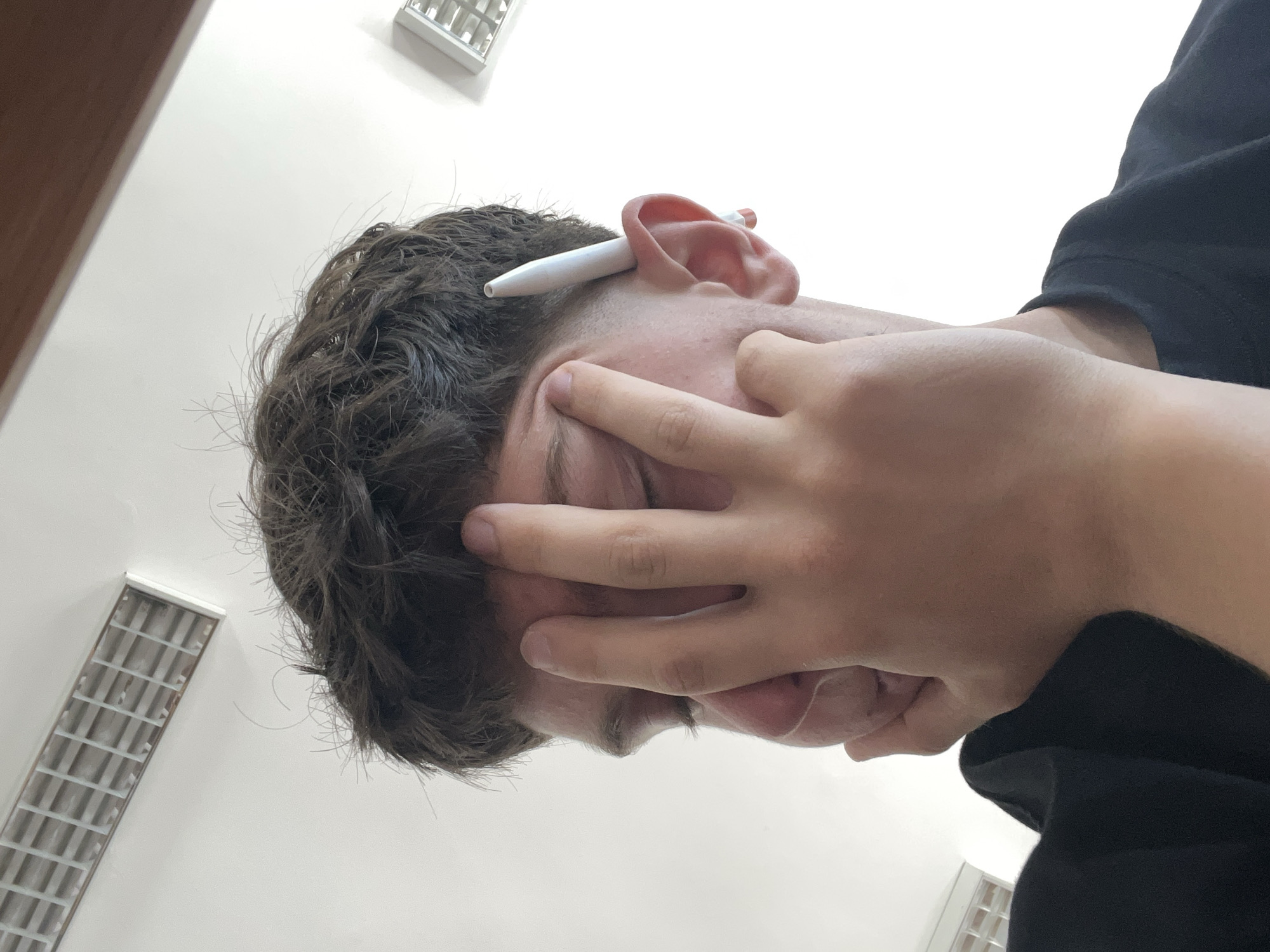 The image shows a young person with their hand covering their face, obscuring most of their features. They have dark, tousled hair and a white stylus is tucked behind their right ear. The person is wearing a black t-shirt. The background consists of a white ceiling with a ventilation grate visible at the top left and another light fixture at the top right. The lighting suggests an indoor environment. The image does not contain any information that would allow for a specific location in the city of Zlatograd, Bulgaria, to be identified.