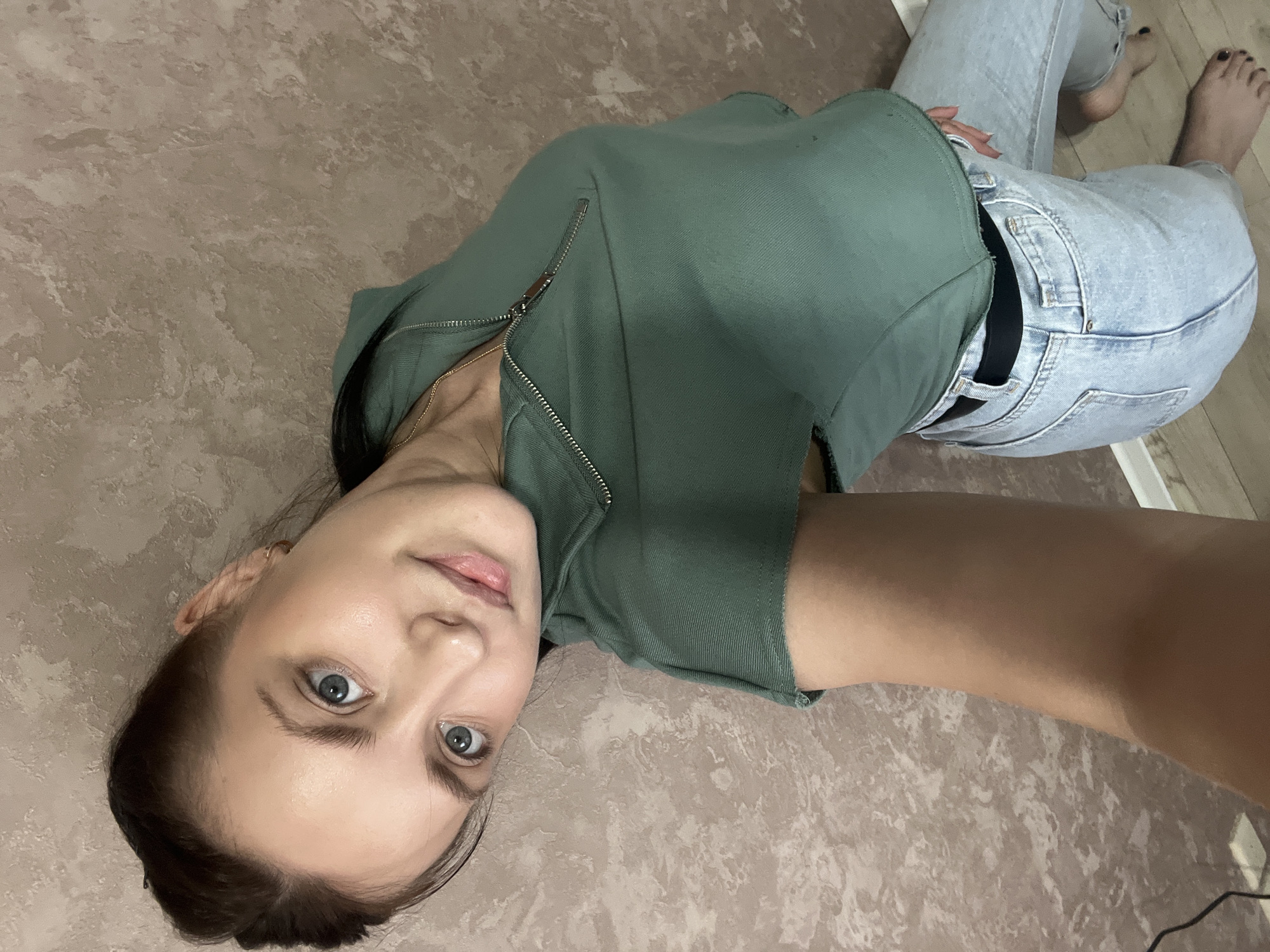 A young woman is lying on a mottled, light brown floor. She is angled with her head toward the lower left corner. She is wearing light-wash denim jeans, a dark-colored belt, and a zipped-up green hoodie. She has dark hair and light eyes. She is barefoot. The location is Марковский, Russia.