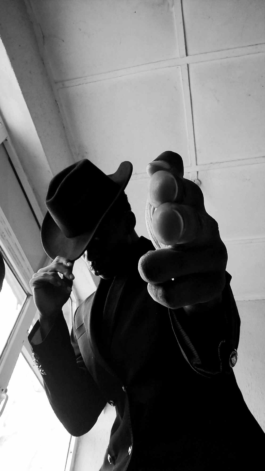 A low-angle, black and white, full-body shot captures a person wearing a hat and a suit jacket, with their right index finger extended towards the camera. The person's face is obscured by shadow, with only a hint of their chin and nose visible. They are standing near a window, which lets in natural light, and the background appears to be a ceiling with a grid pattern. The person's right hand is in sharp focus, with the finger pointing directly at the viewer. The left hand is shown touching the brim of the hat. The overall impression is dramatic and confrontational. This image is located in Sabon Tasha, Nigeria.