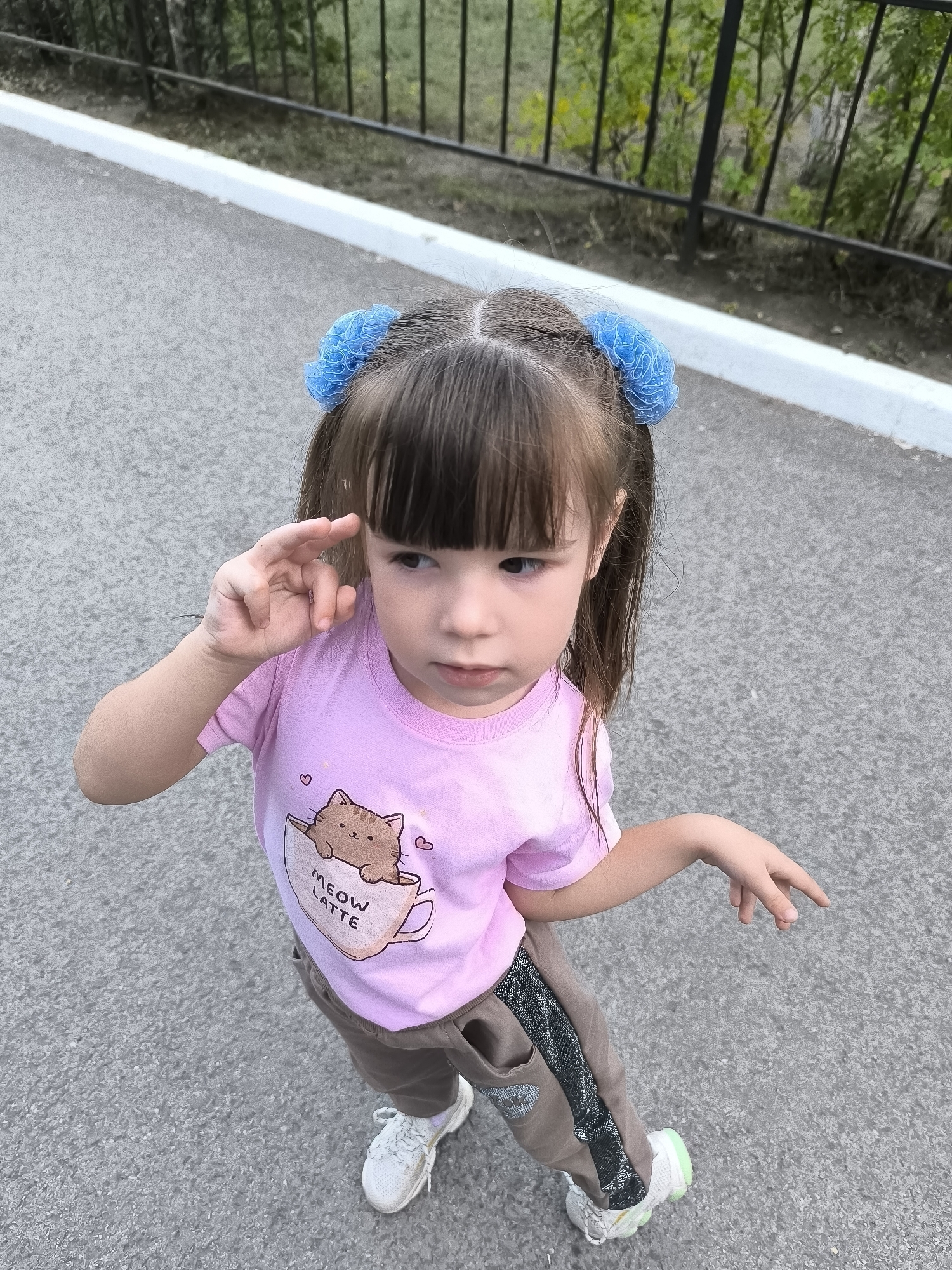 A young girl with brown hair styled in two pigtails with blue scrunchies stands on an asphalt surface. She is wearing a pink t-shirt with a graphic of a cat in a cup and the text "MEOW LATTE," and brown pants with a metallic stripe. She is looking to her right, with her left hand raised to her forehead. The background shows a black metal fence with greenery behind it, and a white curb. The scene appears to be an outdoor location, potentially a park or a pathway.