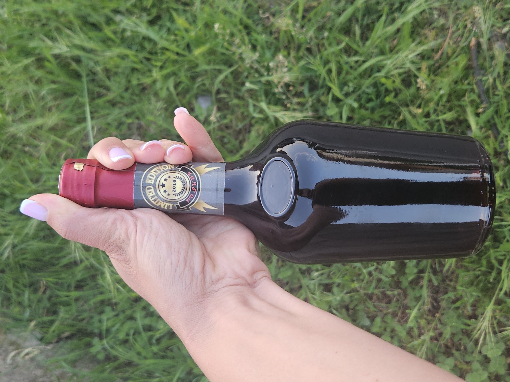 A person's hand holds a dark bottle horizontally against a blurry background of green grass. The hand, which appears to be female, has light skin and manicured fingernails with white tips and pink bases. The bottle is made of dark glass, reflecting the ambient light, and suggests it contains a dark liquid. The neck of the bottle is covered with a red foil or capsule.
A portion of the bottle's label is visible. It is dark with gold or yellow details, featuring stars and text. Clearly visible text includes "LIMITED EDITION" and "2016". Additional partial text that appears to be "ROCK" can also be discerned on the label. The overall setting is outdoors, indicated by the natural light and the grassy background. The image is a close-up shot, focusing on the hand and the bottle.