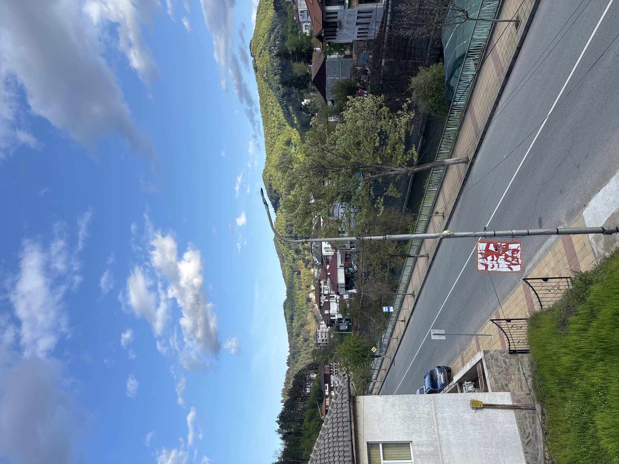The image shows an elevated view of a street in Zlatograd, Bulgaria. A road runs diagonally across the frame, flanked by sidewalks and a low barrier. On the left side of the road, a forested hill rises with interspersed buildings. On the right, a building with a gray roof is partially visible in the foreground. A signpost with a vandalized sign stands beside the street. The sky is blue with scattered clouds. A car is parked beside the building on the right.