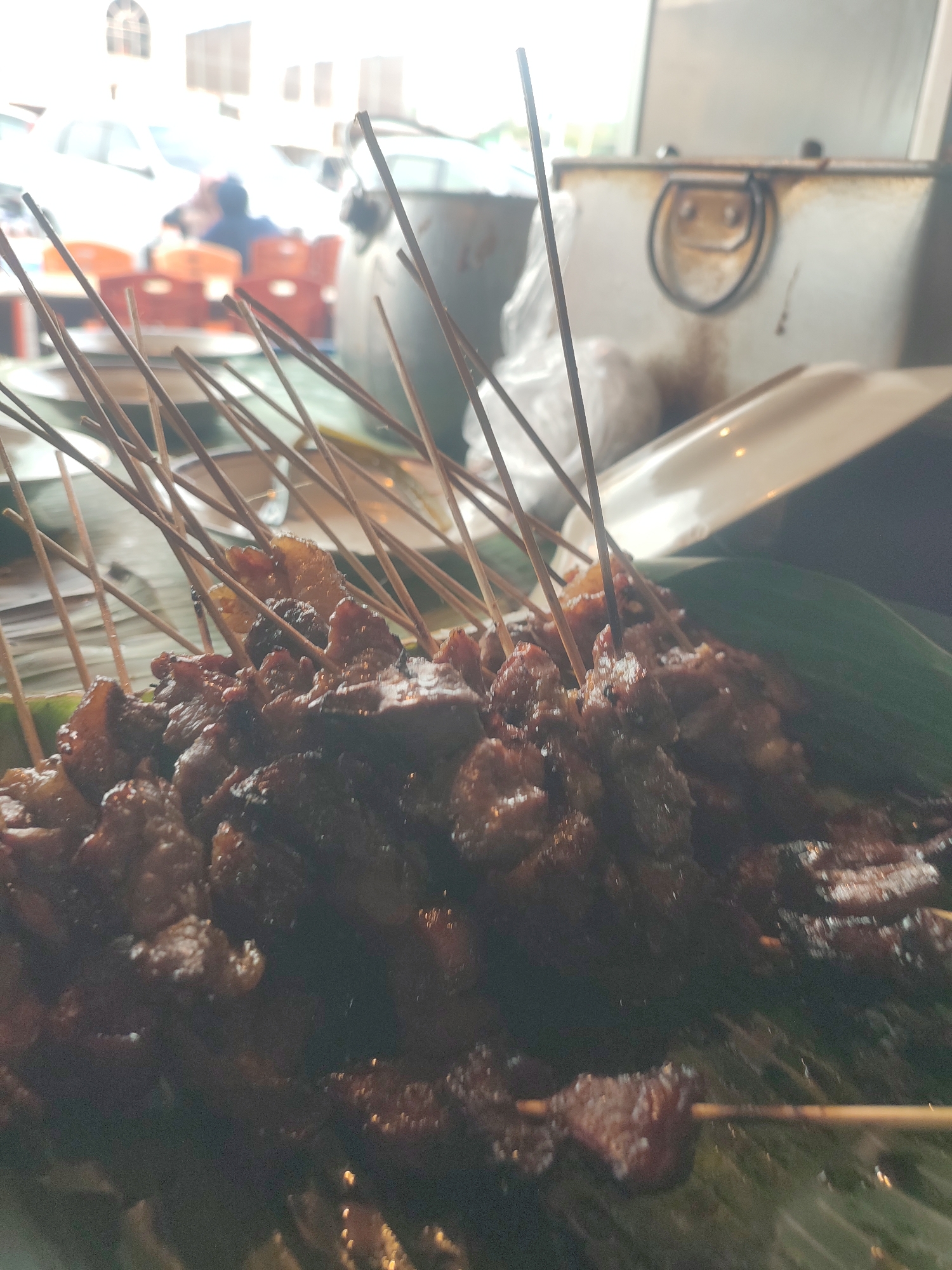 A close-up, slightly blurred photograph captures a tray of satay, likely beef, presented on a bed of banana leaves. The satay skewers are arranged densely, with many sticks protruding upwards. The meat appears grilled and coated in a dark, possibly sweet or savory sauce. In the blurred background, a cooler or metal container with a handle is visible, and beyond that, an outdoor setting with orange chairs and people can be discerned. The lighting suggests an evening or indoor environment. The context provided indicates this scene is in the city of Cotpuuk, Indonesia, a location known for its street food culture, particularly satay.