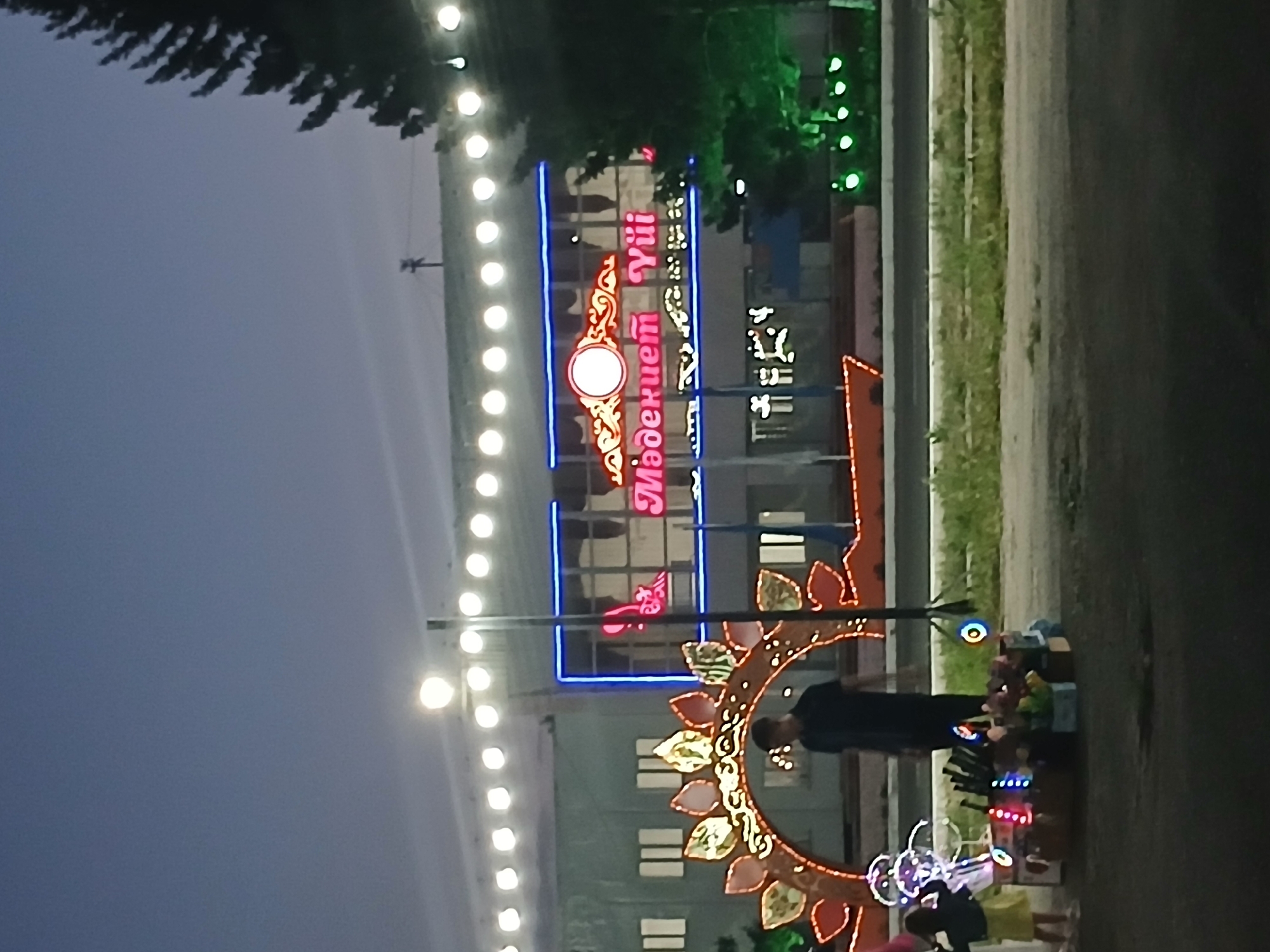 The image depicts an outdoor scene in the city of Makinsk, Kazakhstan, at dusk or early evening. In the background, a building with multiple windows is illuminated by a string of bright white lights along its facade. A neon sign above a section of the building reads "Мәдениет Үиі" (Madeniet Yui). In the foreground, a person in a dark shirt and pants stands near a large, illuminated decorative archway featuring leaf-like motifs. To the left of the archway, a young child in a yellow dress and a darker jacket stands near a cart piled with various items, possibly toys or souvenirs, some of which are also decorated with colorful lights. The ground appears to be asphalt, with a strip of grass and a road visible between the foreground and background. A large evergreen tree stands to the right, also illuminated by green lights. The sky is a deep blue, indicating twilight.