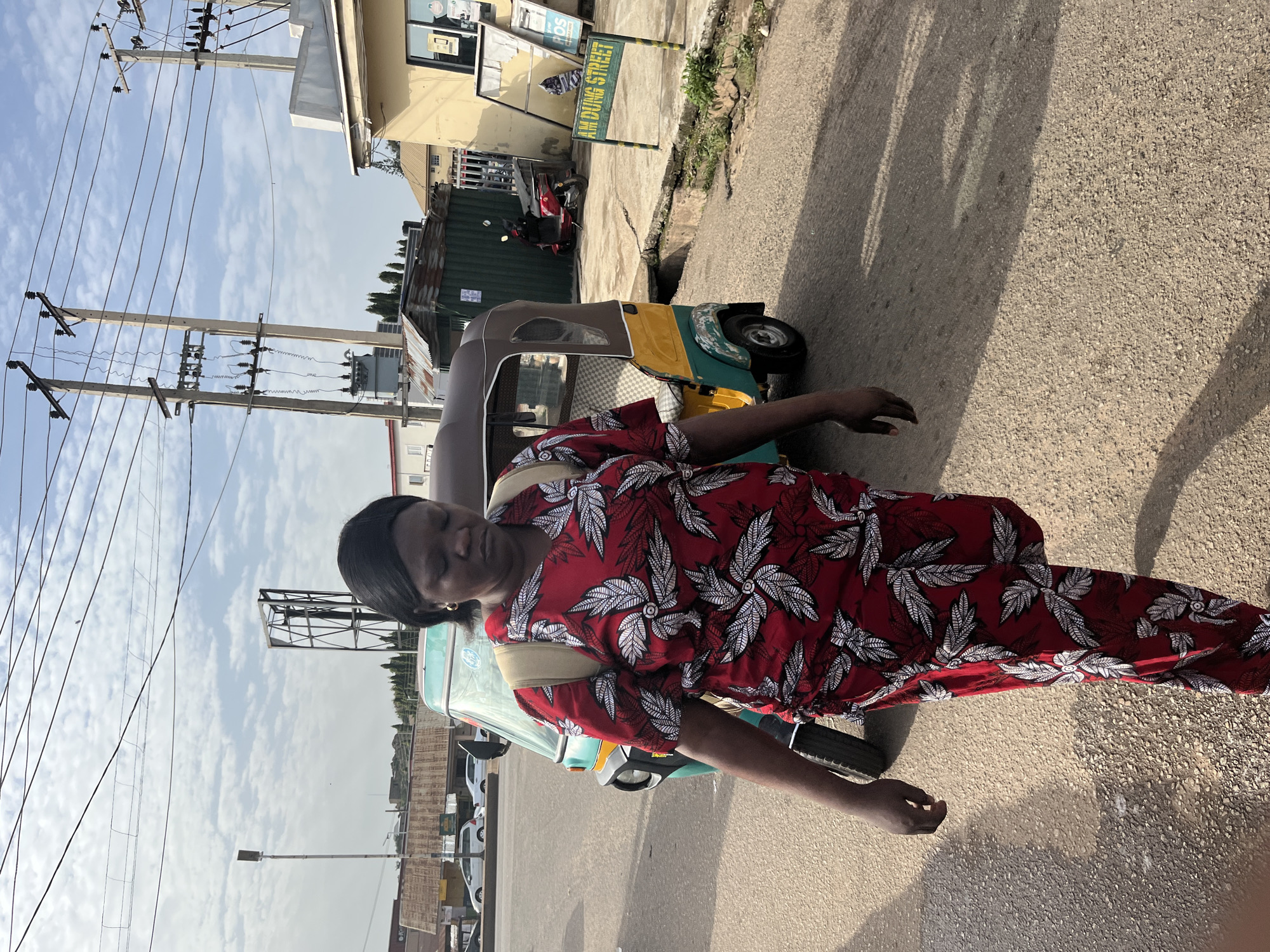 A woman walks on a street in Jos, Nigeria, in front of a three-wheeled taxi. She wears a red dress with a white leaf pattern and a beige backpack. The street is paved with asphalt and gravel. Behind her, a yellow and green three-wheeled taxi is parked. In the background, there are buildings, utility poles, power lines, and a sign that reads "AM DUNG STREET." The sky is partly cloudy.